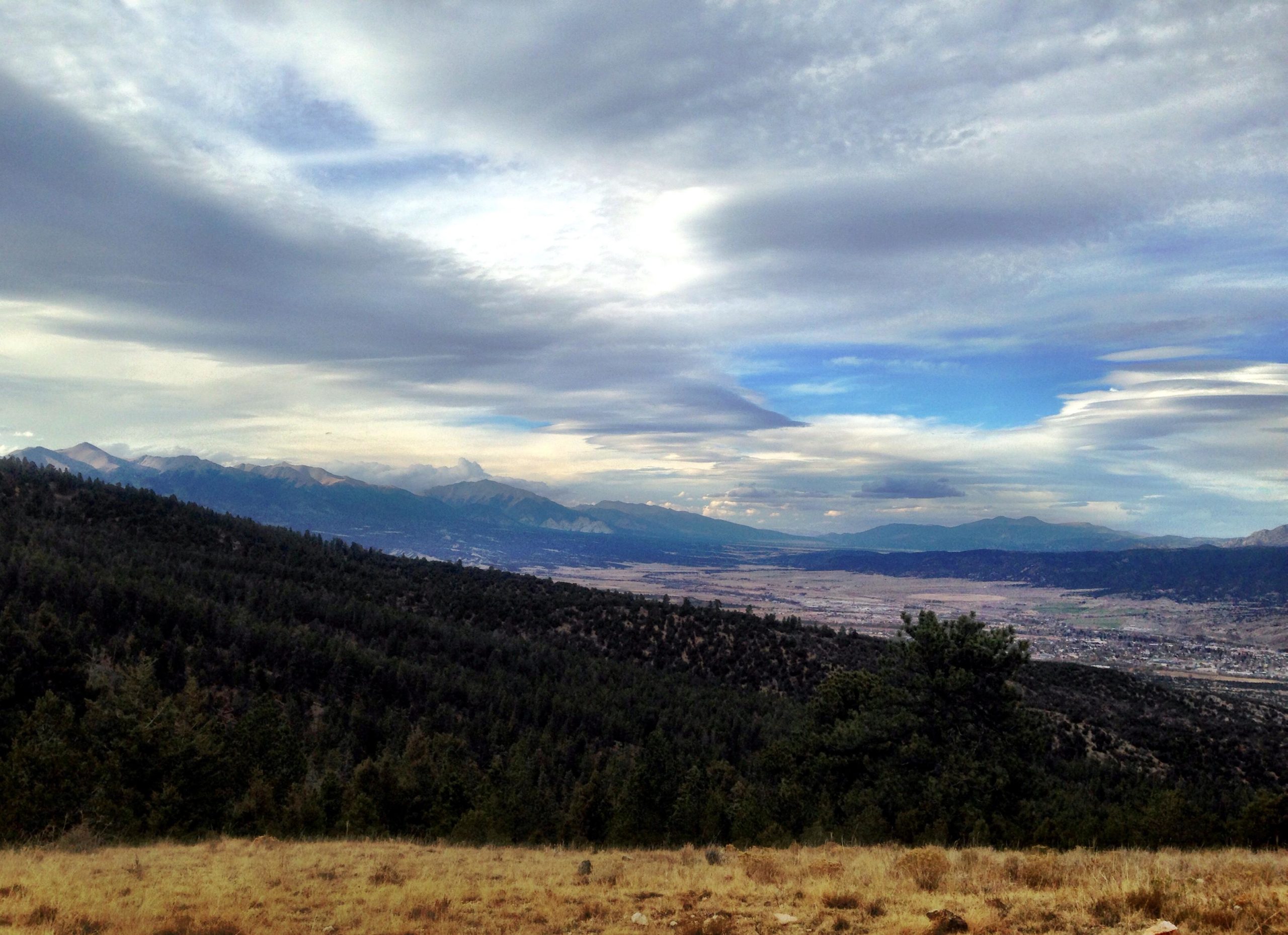 A panoramic view of a mountainous landscape under a cloudy sky, featuring rolling hills covered in greenery and a valley below. The mountains in the distance have a mix of rocky peaks and gentle slopes, with patches of snow visible at the highest points. The sky is adorned with various cloud formations, casting soft light over the scene. Rainbow Trail: Methodist Mountain Thd to Bear Creek Thd mountain bike trail.
