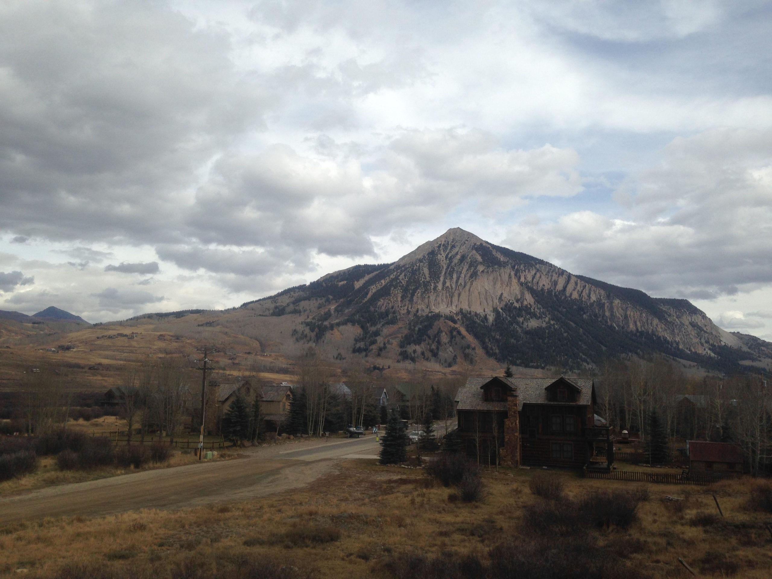 A scenic view of a mountain landscape with a prominent peak in the background, surrounded by rolling hills and sparse tree coverage. In the foreground, a dirt road leads toward a rustic wooden building with a few trees nearby. The sky is partly cloudy, adding depth to the overall natural scene. Upper Lower Loop mountain bike trail.