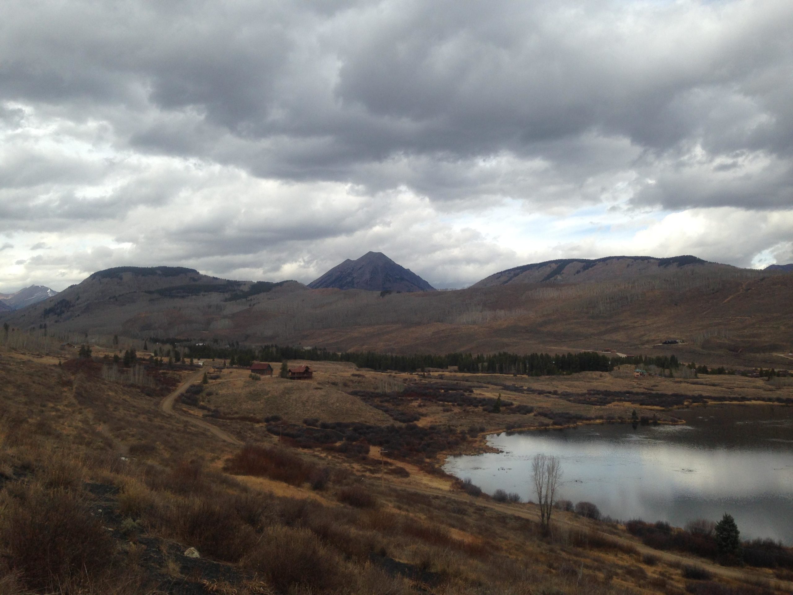 A scenic view of a mountainous landscape under a cloudy sky. The foreground features a calm pond reflecting the overcast sky, surrounded by sparse vegetation. In the background, rolling hills and peaks are visible, with some areas showing patches of snow. A dirt road winds through the landscape, leading to a few rustic buildings nestled in the valley. Upper Lower Loop mountain bike trail.