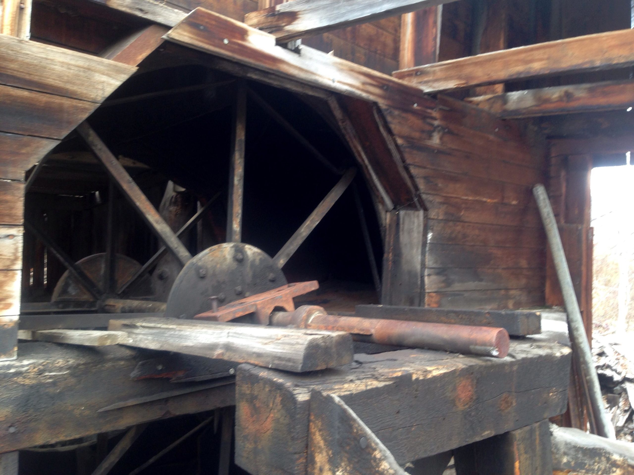 An abandoned wooden structure with a large circular wheel and various tools on a workbench. The wood is aged and weathered, showcasing a rustic, industrial atmosphere. The scene captures elements of decay and history, with a dark interior visible through the opening of the building. Upper Lower Loop mountain bike trail.