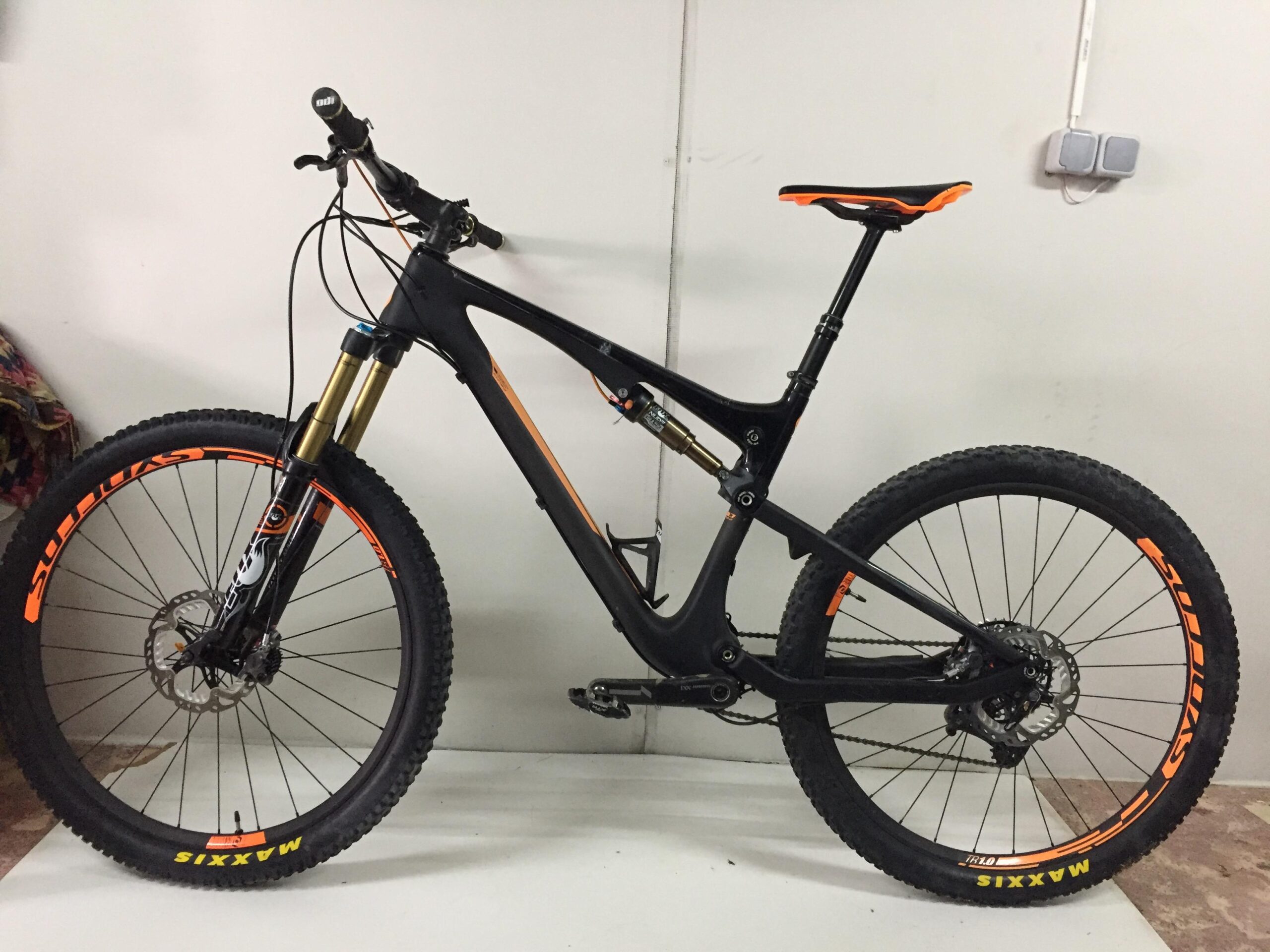 Scott Genius 700 Tuned Bike: A black and orange mountain bike is displayed against a white wall. The bike features thick tires with bold orange lettering, a suspension fork, and a sleek frame design. The seat is raised, and the handlebars are angled slightly, showcasing the bike's ready-to-ride appearance.