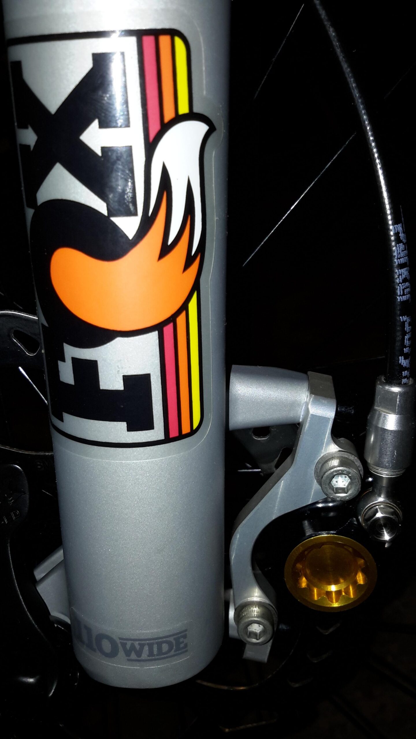 Orbea Loki: Close-up image of a bicycle's fork featuring a vibrant FOX Racing Shox logo, with a stylized flame design and color stripes. The fork is mounted to a brake assembly, showcasing metallic components, a golden hydraulic feature, and the label "110 WIDE" on the lower part of the fork. The background is dark, enhancing the visibility of the details.