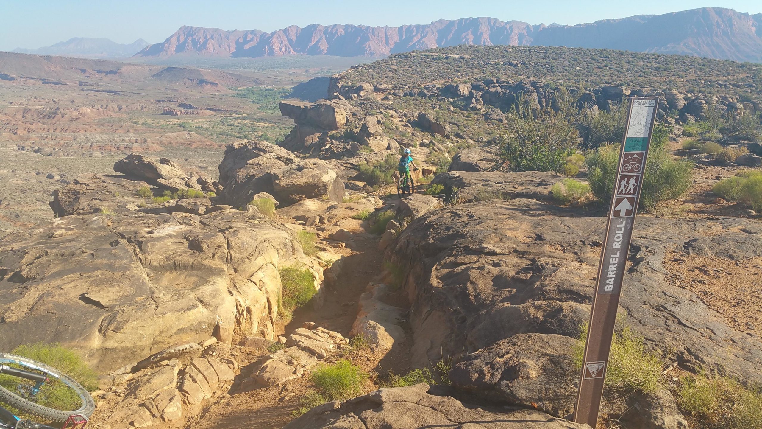A scenic view of a rocky landscape with a distant range of mountains under a clear sky. In the foreground, a trail sign labeled "Barrel Roll" is visible, indicating a path for biking and hiking. A person in a teal outfit is seen standing on the trail with a bicycle, overlooking the terrain. Green shrubs and grasses grow among the rocks, adding to the natural beauty of the area. Barrel Roll mountain bike trail.