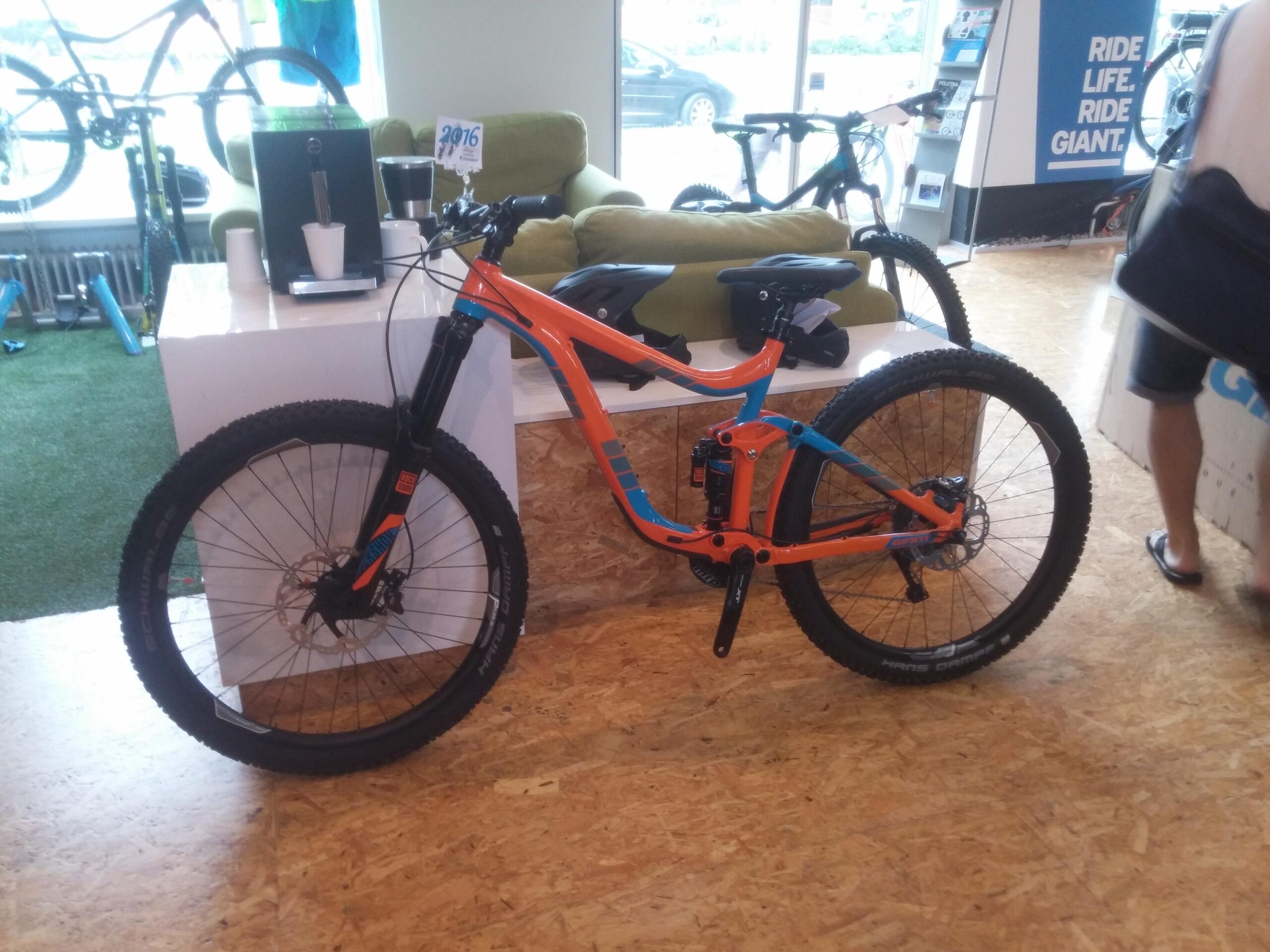 Giant Reign: An orange and blue mountain bike displayed in a shop with a modern interior. There is a coffee machine and cups on the counter behind the bike, and a green couch is visible in the background. Other bikes are partially visible in the background, along with a person in shorts and sandals walking by.
