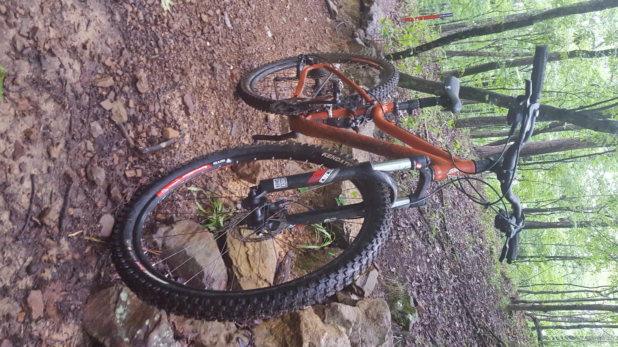Salsa Big Mama SRAM: A mountain bike resting on a rocky, muddy trail in a lush green forest. The bicycle is primarily orange with black tires and components, positioned on uneven terrain surrounded by trees and foliage.