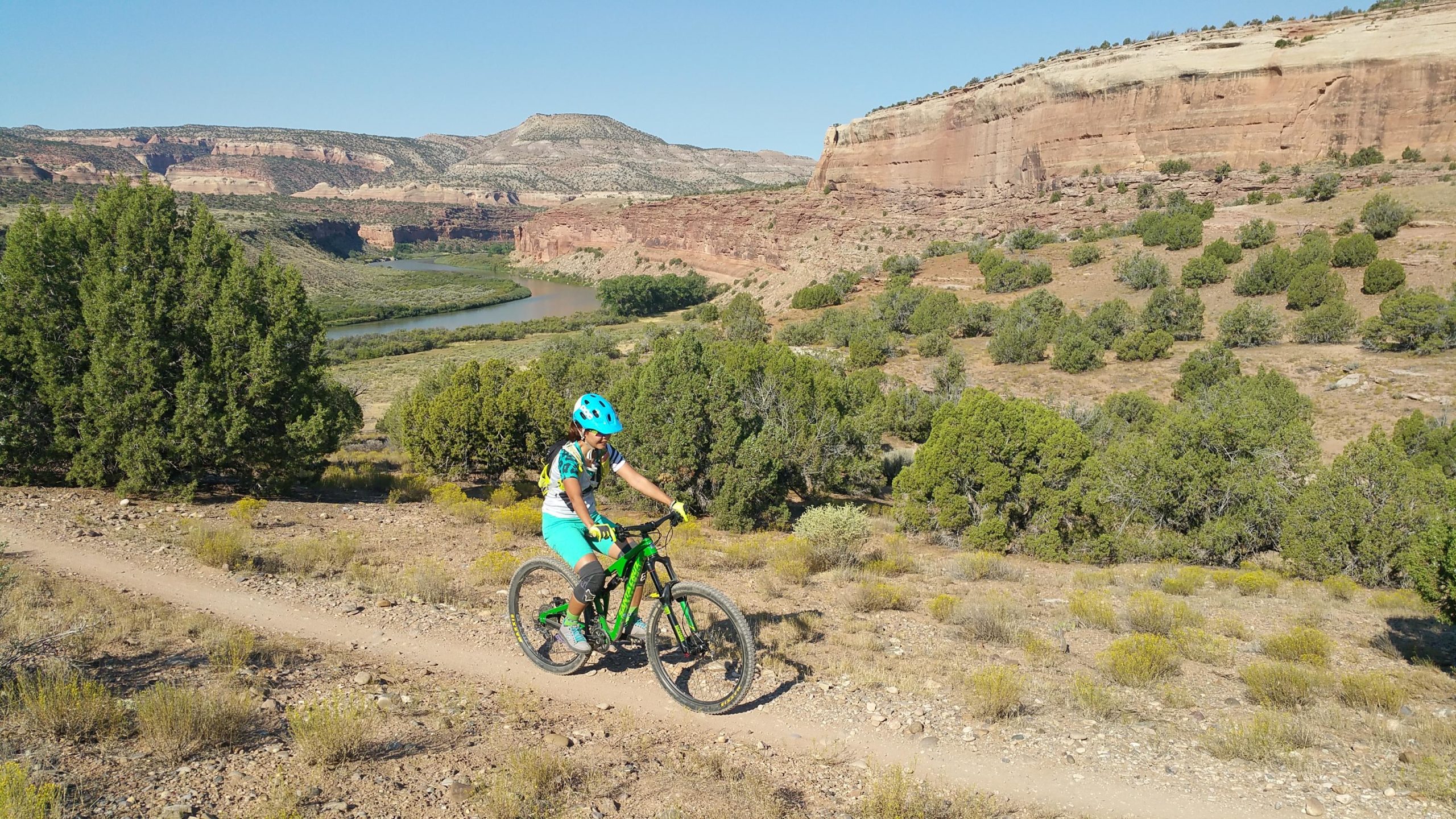 A mountain biker riding along a dirt trail in a scenic landscape, surrounded by greenery and rocky cliffs. In the background, a winding river flows through the valley under a clear blue sky. Mary's Loop / Horsethief Bench mountain bike trail.