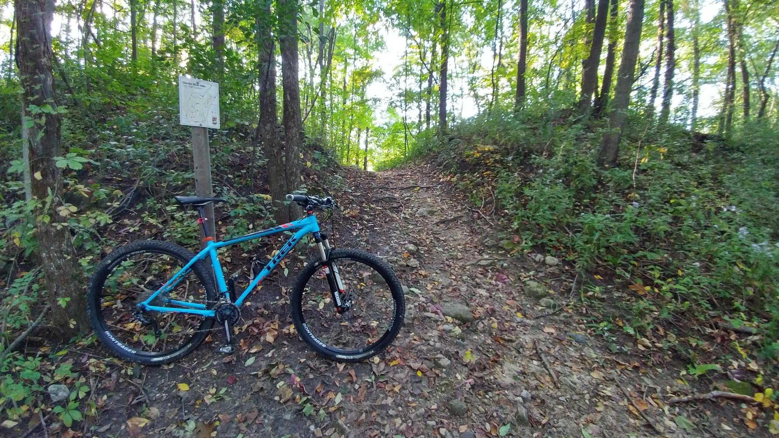 Trek X-Caliber 9: A blue mountain bike parked next to a dirt trail surrounded by trees and foliage, with a trail map sign in the background. The ground is covered in leaves, indicating a natural, wooded area ideal for biking.