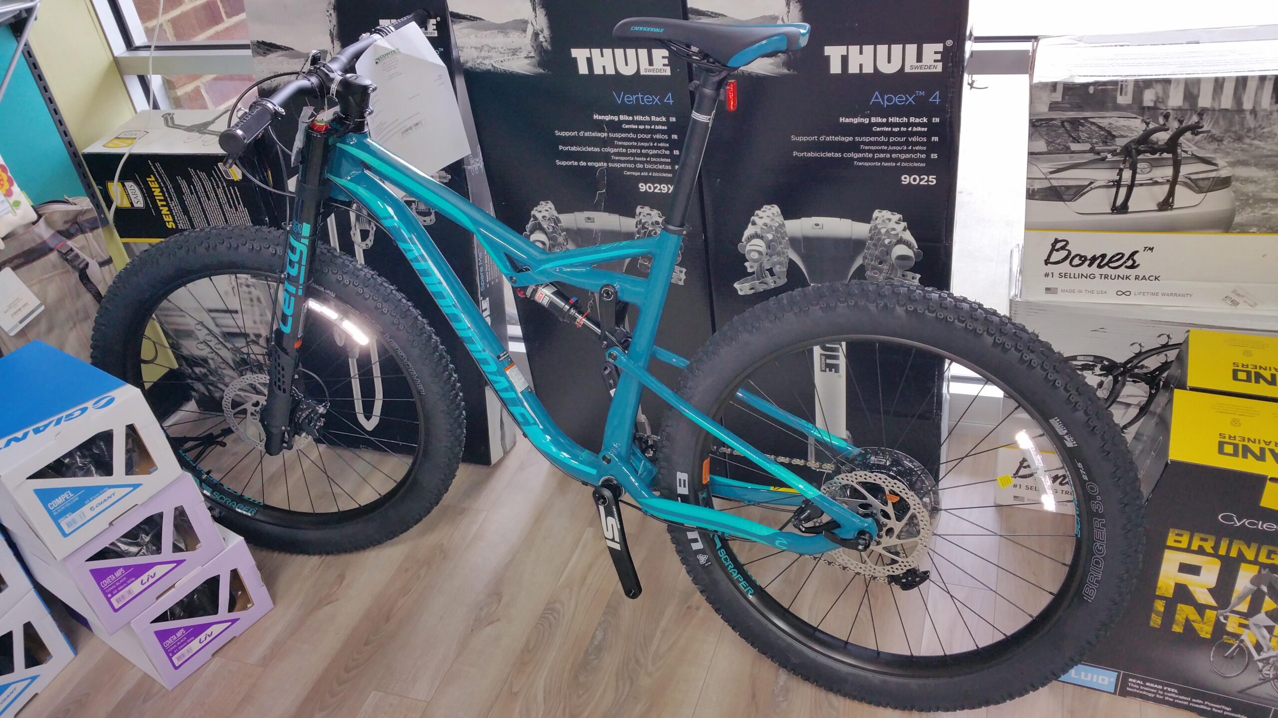 Cannondale Bad Habit: A teal mountain bike with fat tires is displayed in a store surrounded by boxes of bike accessories. The bike features a front suspension fork, disc brakes, and a prominent chainring. Various bike rack products are visible in the background. The flooring is wood-like, adding to the indoor shopping environment.