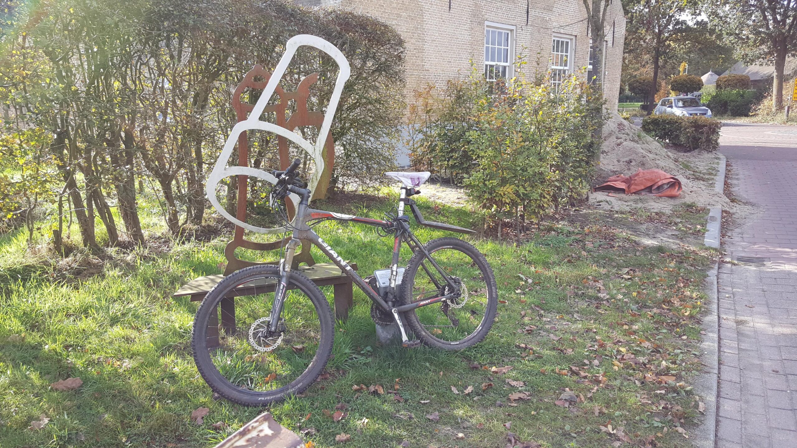 Giant Talon 27.5: A mountain bike leaning against a brown wooden bench, with a large white sculpture resembling a chair positioned behind it. Surrounding the scene are bushes and trees, with fallen leaves scattered on the ground. In the background, a sandy area and a parked car are visible.