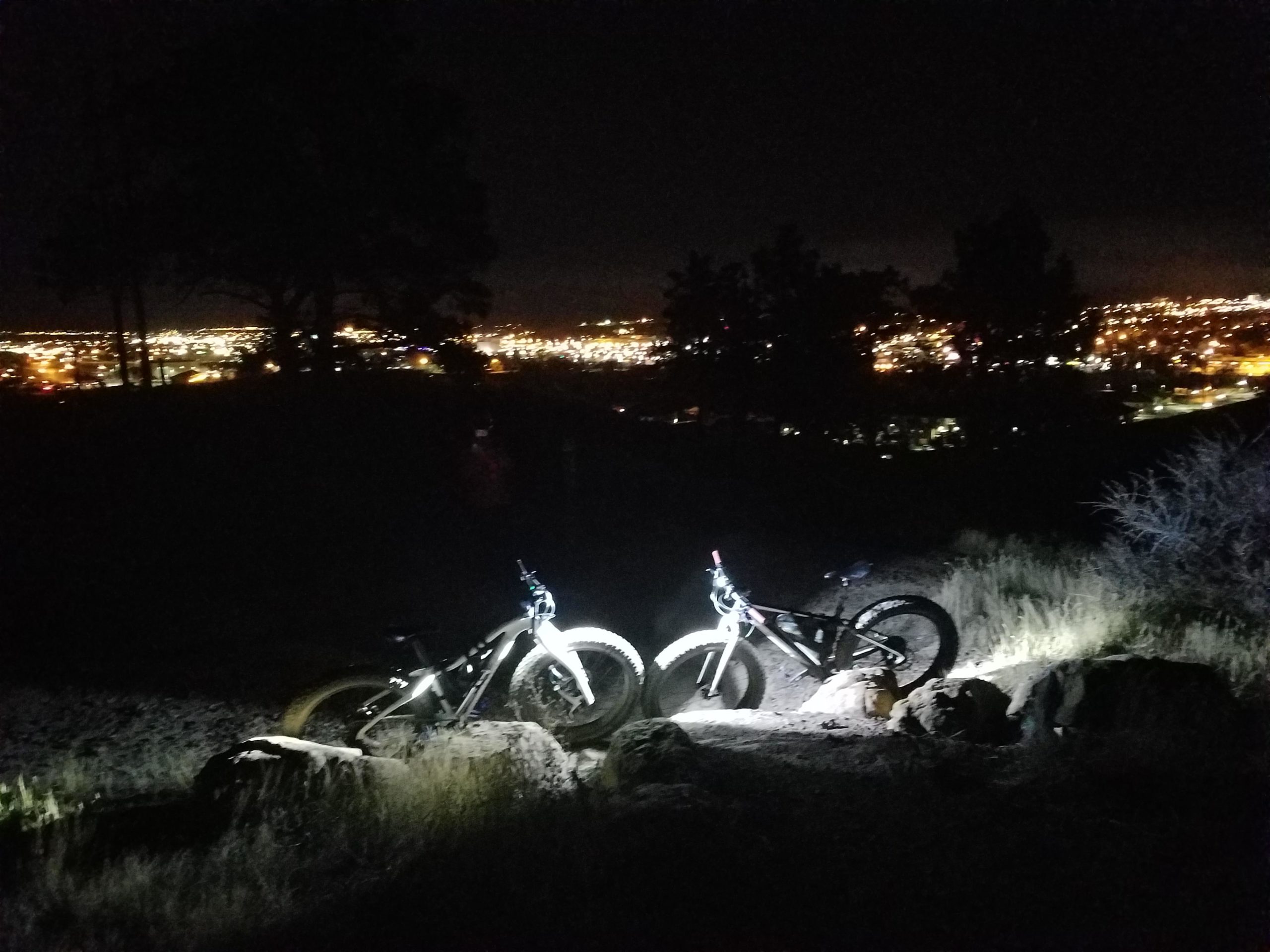 Two fat bikes are illuminated by their lights, parked on rocky terrain at night. In the background, a city skyline is visible, twinkling with lights against the dark sky. Silhouetted trees frame the scene, enhancing the nighttime ambiance. HLMP mountain bike trail.