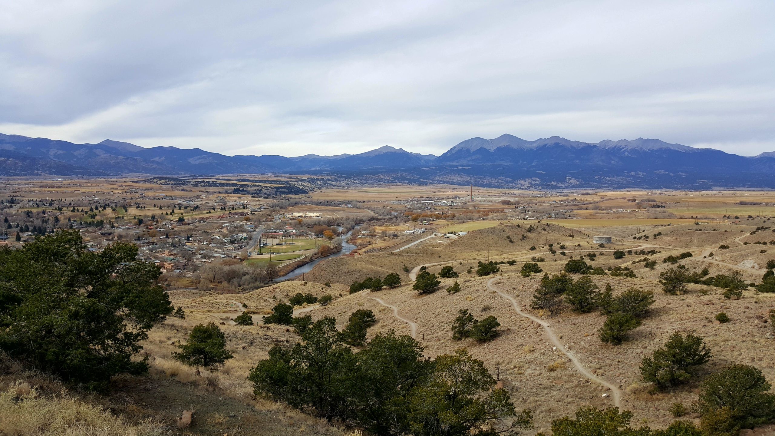 A panoramic view of a valley surrounded by mountains, featuring patches of trees and a winding river flowing through a small town. The landscape showcases rolling hills in the foreground, with softly undulating terrain leading to the distant mountains under a cloudy sky. Arkansas Hills mountain bike trail.
