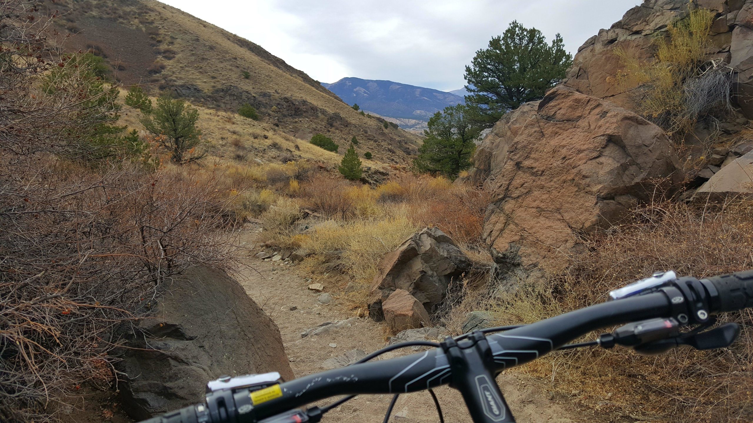A view of a mountain biking trail, captured from the perspective of a cyclist. The image shows a narrow dirt path winding through rocky terrain and dry bushes, with hills and mountains in the background under a cloudy sky. The handlebars of the bike are visible in the foreground. Arkansas Hills mountain bike trail.