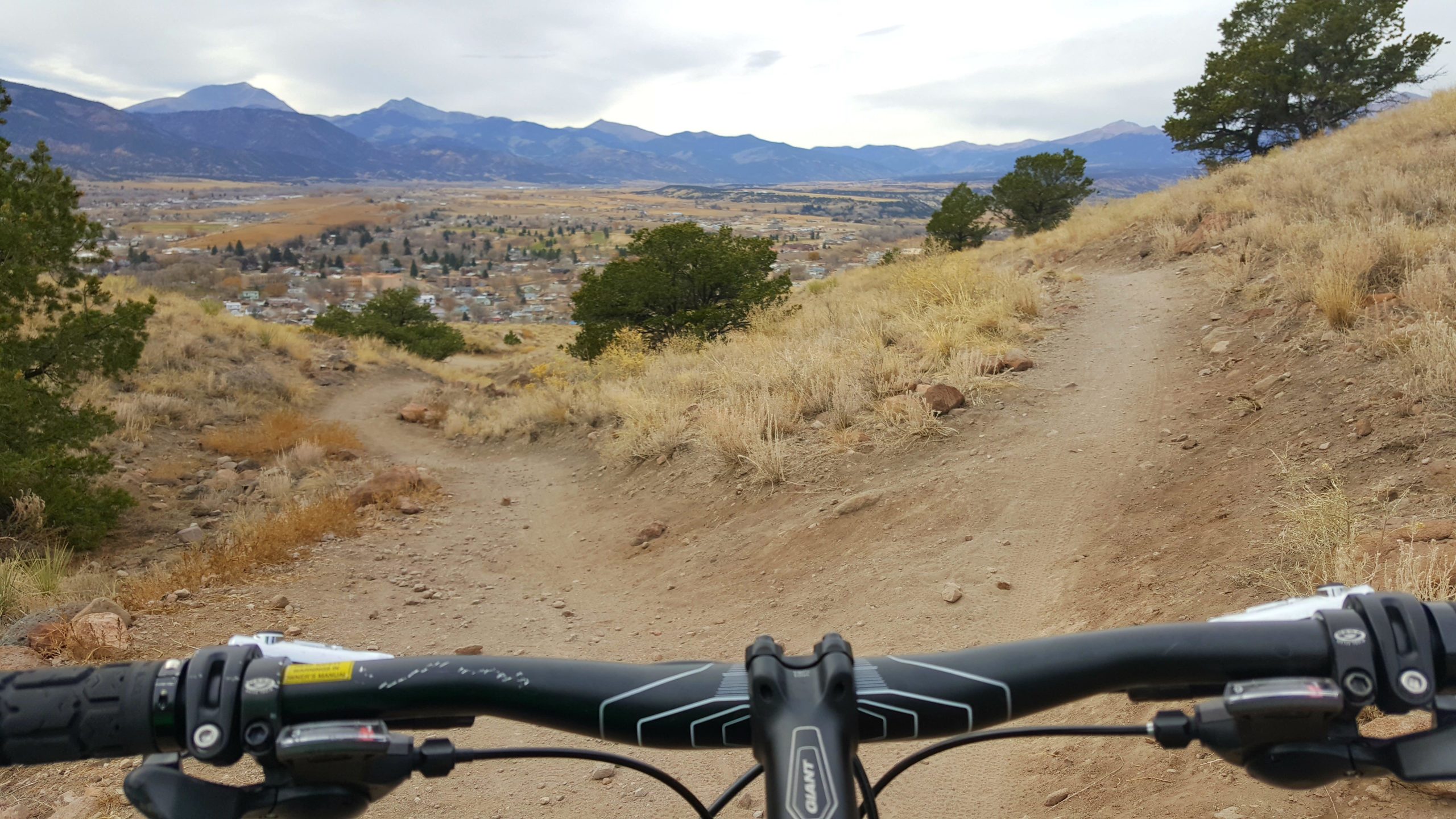 A view from the handlebars of a mountain bike, looking down a dirt trail surrounded by tall grass and rocky terrain, with mountains and a valley town visible in the background under a cloudy sky. Arkansas Hills mountain bike trail.