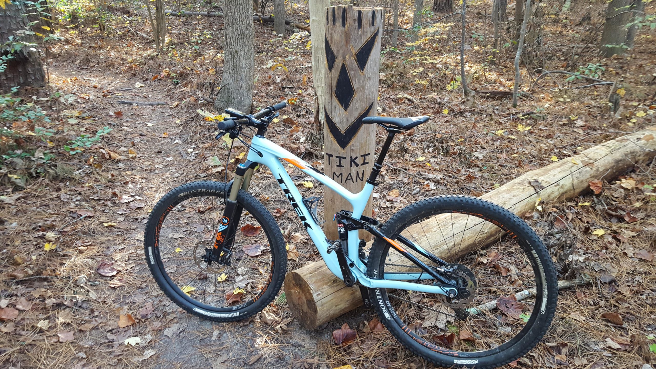 A mountain bike leaning against a wooden totem labeled "TIKI MAN" on a wooded trail, surrounded by fallen leaves and trees. Governor's Creek mountain bike trail.