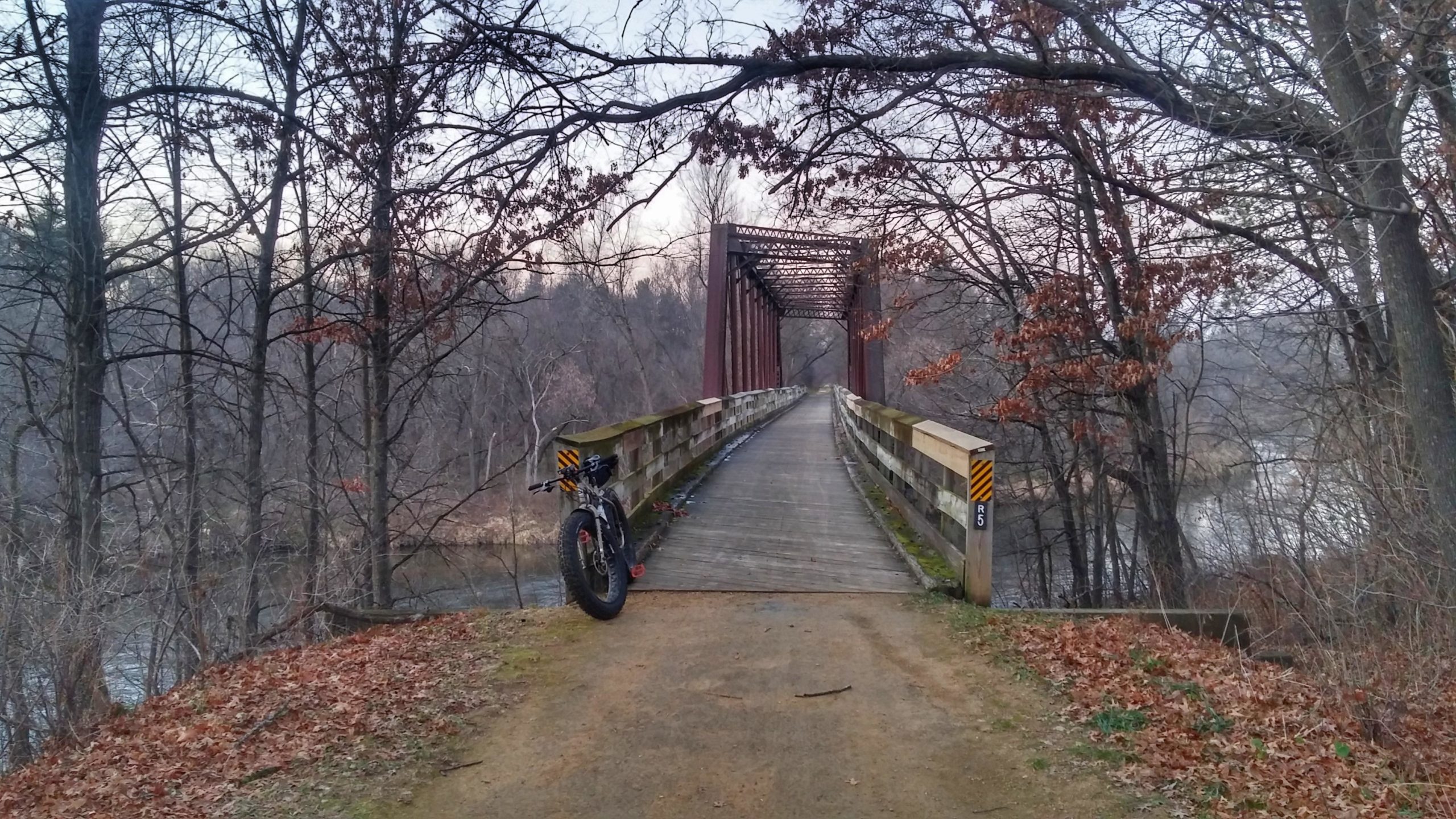 A wooden bridge spans a calm river surrounded by bare trees, with autumn leaves scattered on the ground. A bicycle is leaning against the bridge railing, leading the viewer