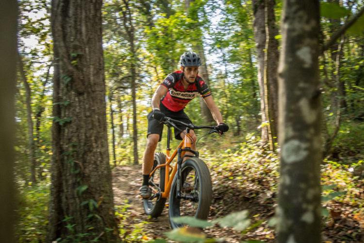 A cyclist in a red and black jersey rides a fat bike along a winding trail in a wooded area. Sunlight filters through the trees, highlighting the natural surroundings as the rider navigates the terrain.