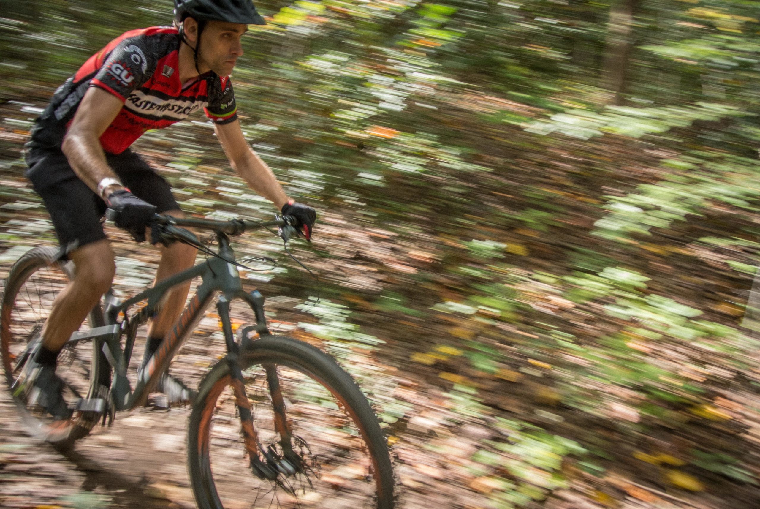 A cyclist wearing a red and black jersey rides a mountain bike along a dirt trail in a wooded area, with motion blur emphasizing speed. The ground is covered in leaves and small plants, capturing the essence of outdoor biking. USNWC mountain bike trail.