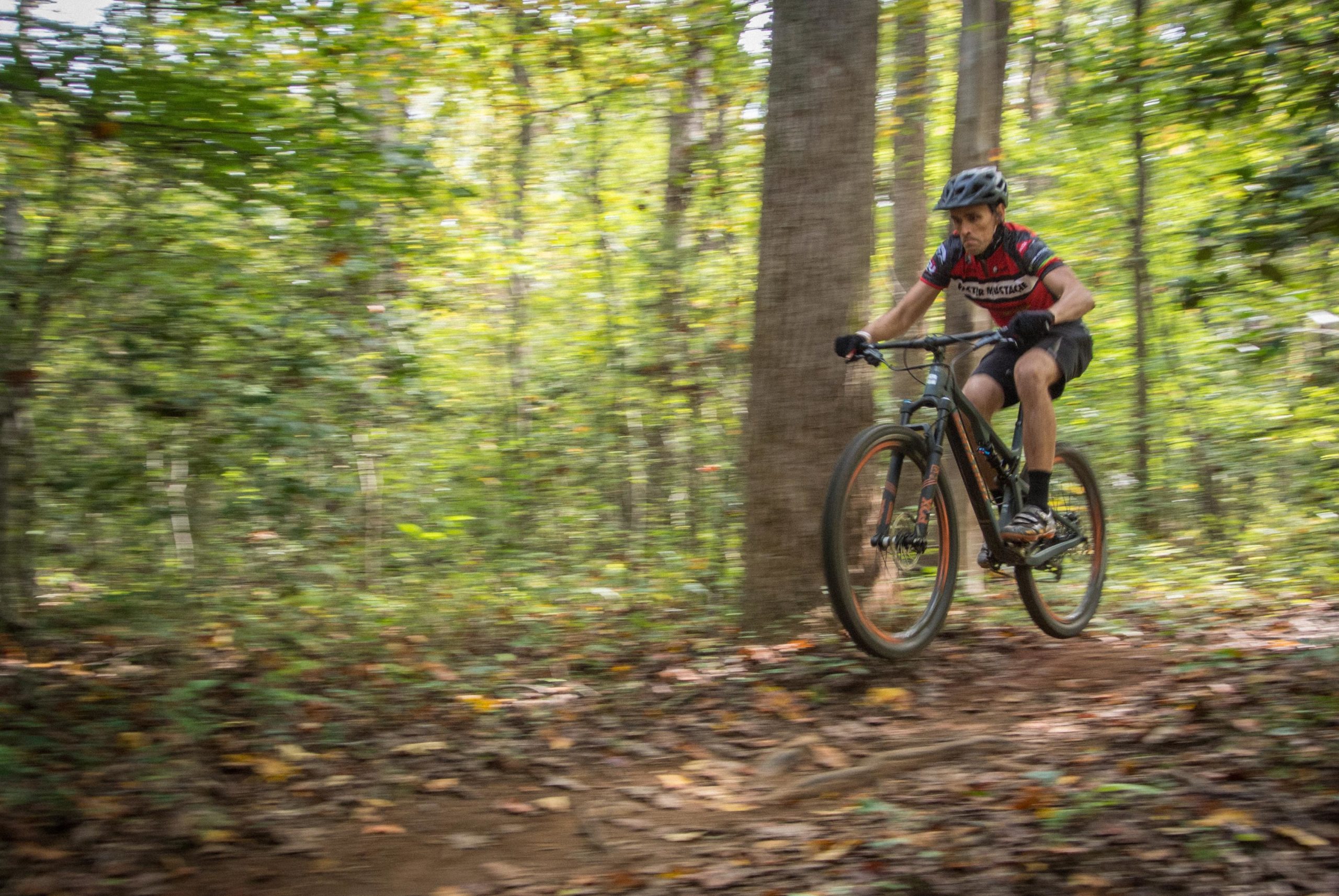 A mountain biker navigating a winding trail through a lush, green forest, captured in motion with blurred background to emphasize speed and action. The rider is wearing a helmet and a colorful cycling jersey, focused on the path ahead. USNWC mountain bike trail.