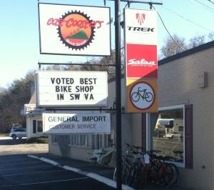 Image of a bike shop storefront featuring a sign that reads "Voted Best Bike Shop in SW VA." The shop displays logos for Trek and Salsa, along with bicycles parked outside. A "General Import" sign indicates customer service. The setting is in a rural area with trees in the background.