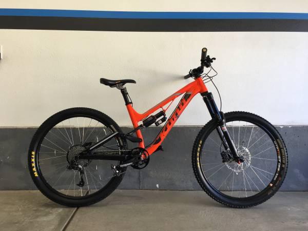 Kona Process 153: Alt text: A mountain bike with a vibrant orange and black frame, featuring wide tires and a suspension fork, is leaning against a gray wall with blue and black stripes.