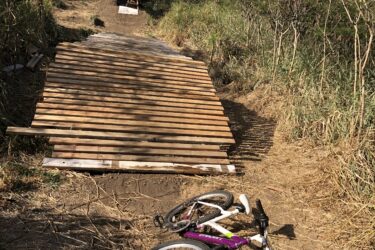 A dirt path leading to a wooden ramp is surrounded by tall grass and shrubs. A white and purple bicycle is lying on its side near the bottom of the ramp, suggesting that it has recently been used. The sky above is clear with some scattered clouds. Kalaheo Hillside mountain bike trail.