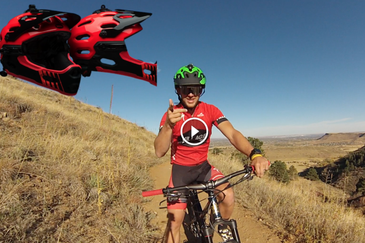 A mountain biker wearing a red jersey and a green helmet poses on a dirt trail, pointing towards the camera. In the background, a scenic view of hills and valleys is visible under a clear blue sky. Three red mountain bike helmets are displayed in the top left corner.