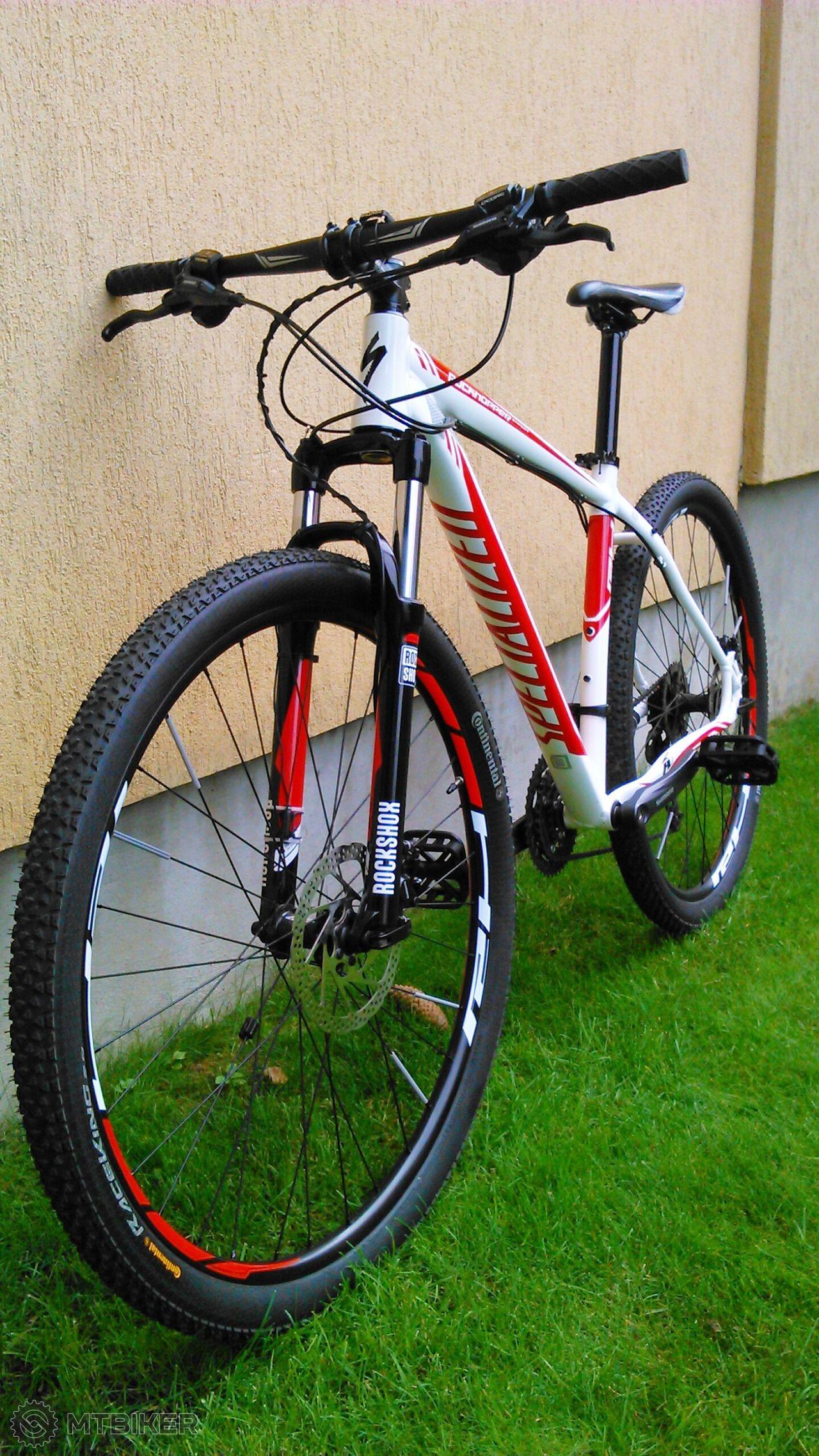 Specialized Rockhopper Expert Disc 29er: A white and red mountain bike leaning against a light-colored wall, featuring thick, treaded tires and a front suspension. The bike's handlebars and seat are visible, positioned above a grassy area.
