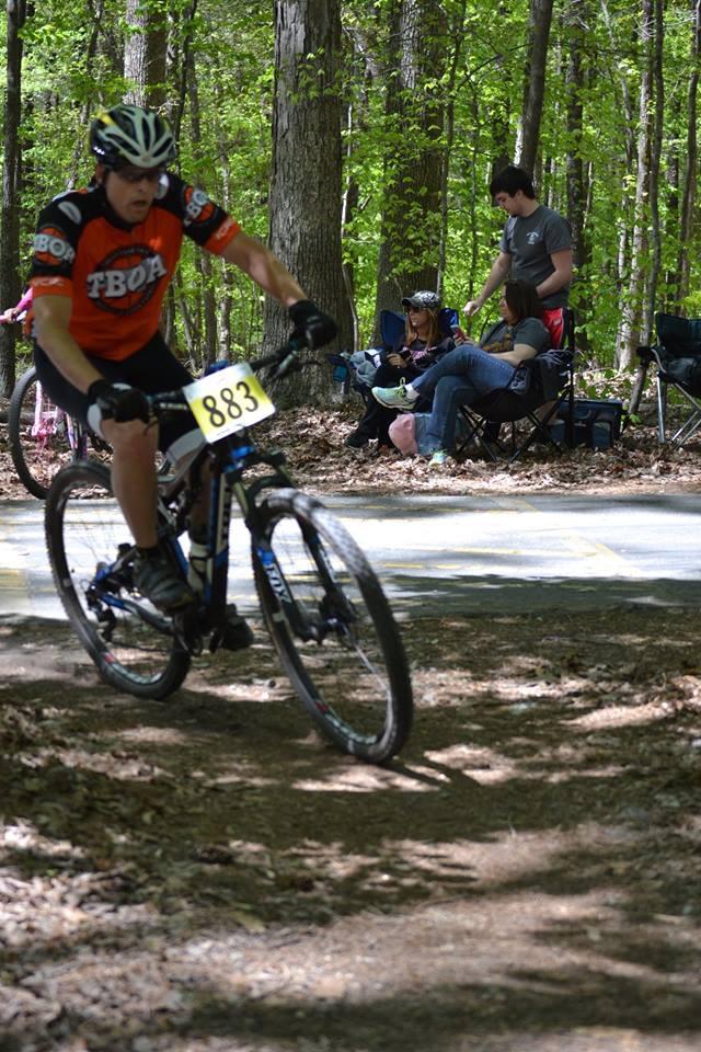 Trek Superfly 100 Elite carbon: A mountain biker in an orange jersey with the number 883 rides along a dirt path through a wooded area. In the background, a small group of people relax in camping chairs, enjoying the scenery. Sunlight filters through the trees, highlighting the lush greenery of the surroundings.