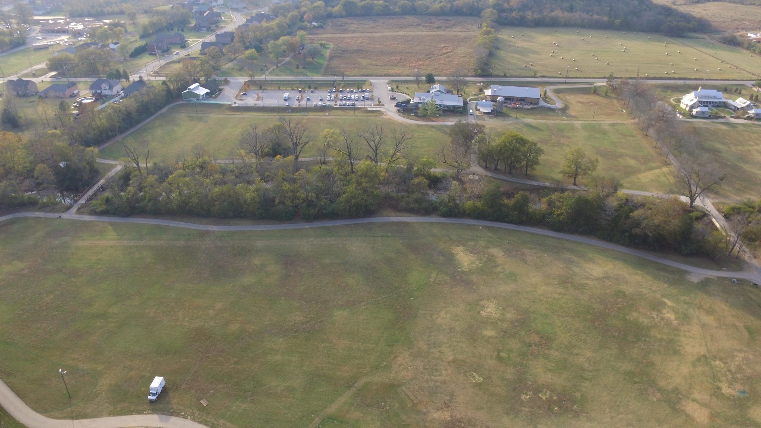 Aerial view of a rural area featuring open green fields, winding paths, and clusters of trees. In the background, there are residential houses, a parking lot filled with vehicles, and several structures, including a barn and another building. The landscape shows a mixture of grassy areas and patches of brown, indicating varied vegetation. The Trails At Fontanel mountain bike trail.