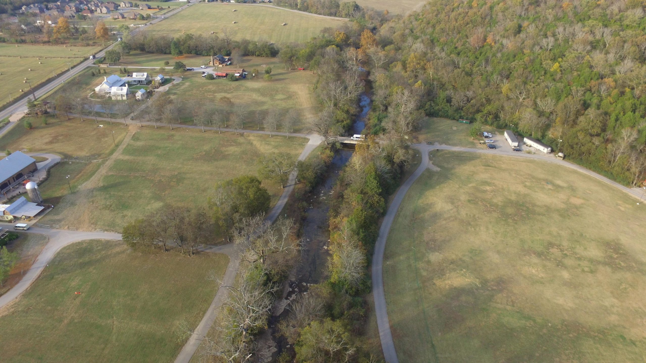 Aerial view of a rural landscape featuring a winding creek surrounded by grassy fields, winding roads, and several houses. The scene includes patches of wooded areas and some farm structures, highlighting the natural beauty of the countryside. The Trails At Fontanel mountain bike trail.