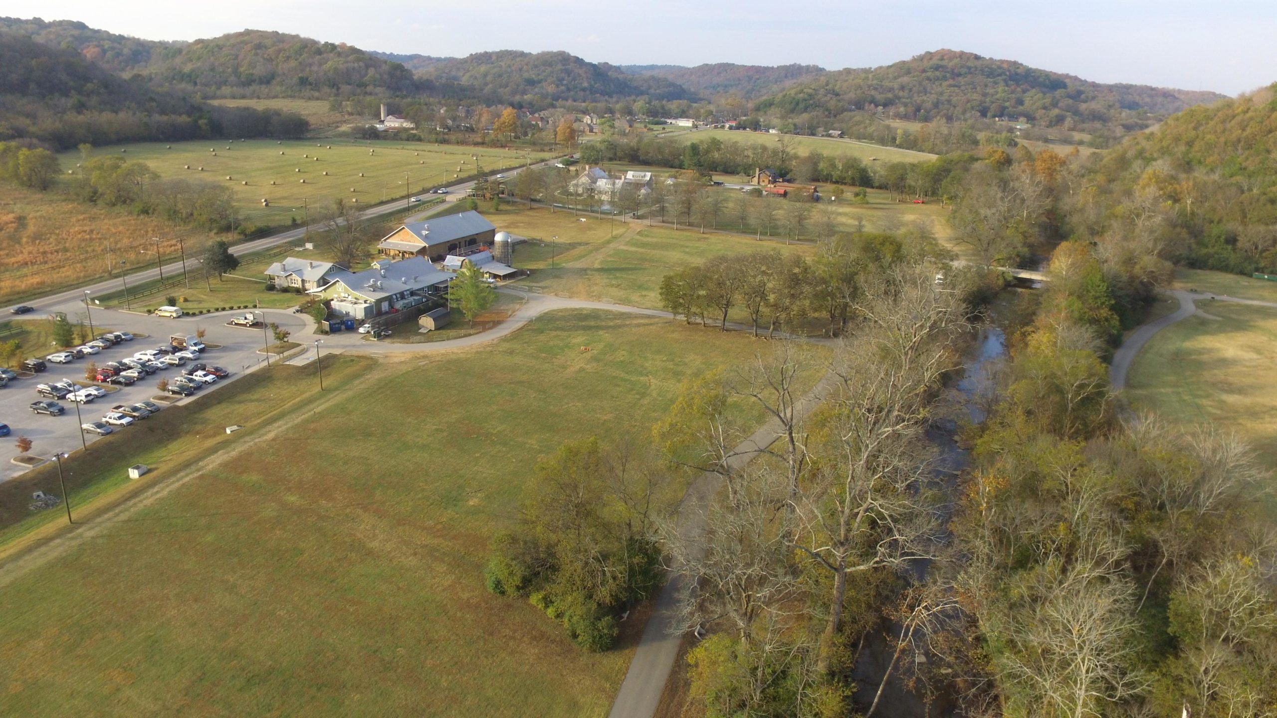 Aerial view of a rural landscape featuring green fields, a winding creek, and a collection of buildings, including a light-colored structure and a larger building with a barn-like appearance. The area includes a parking lot with several cars, a roadway, and hills in the background adorned with autumn foliage. The Trails At Fontanel mountain bike trail.