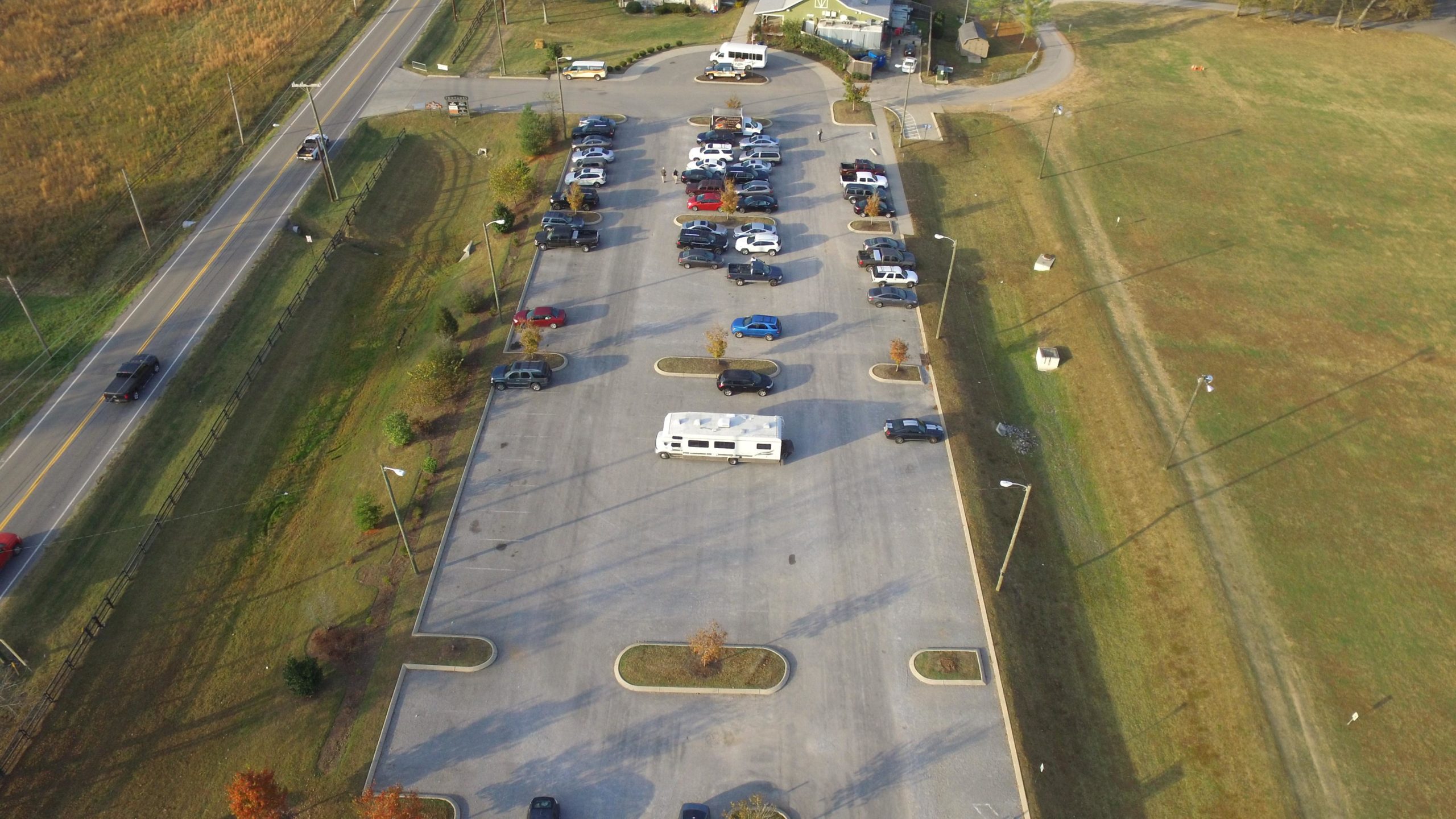 Aerial view of a large parking lot with various vehicles, including an RV, surrounded by grassy areas and a road. The parking lot is partially occupied, with neatly arranged parking spaces visible. In the background, a building and additional vehicles can be seen near the entrance. The Trails At Fontanel mountain bike trail.