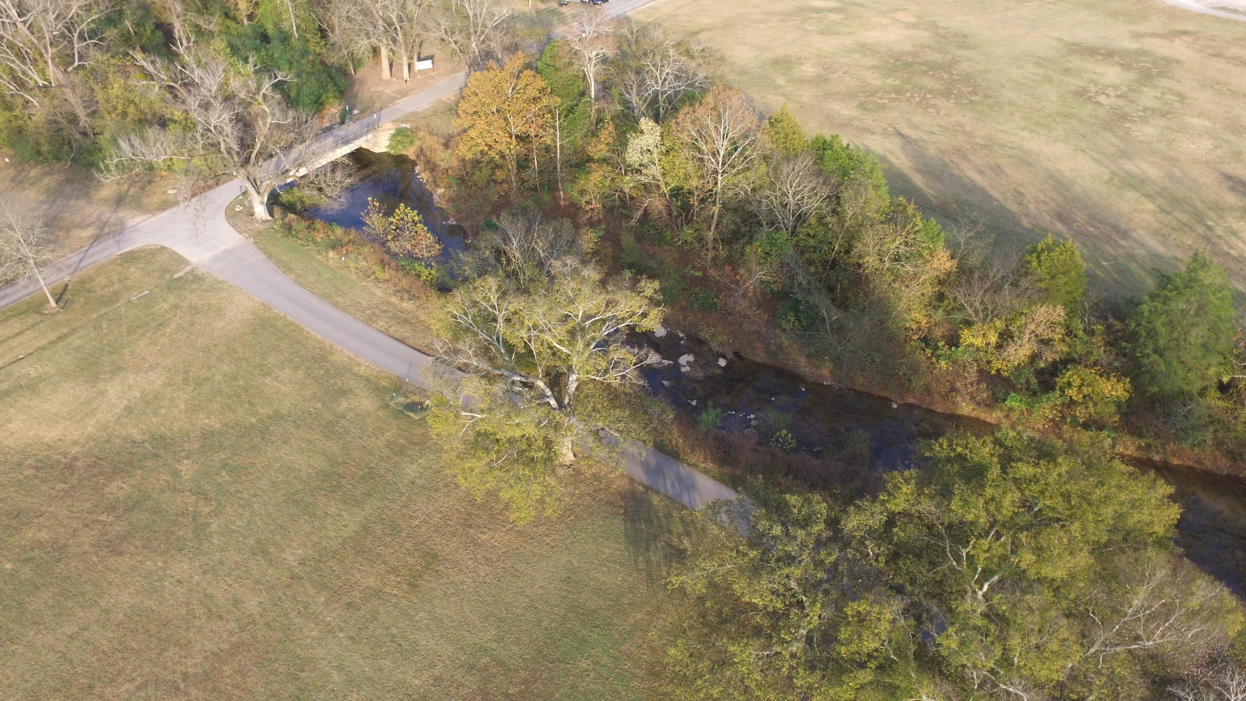Aerial view of a park featuring a winding path alongside a stream, with a small bridge crossing over. Surrounding the stream are trees displaying autumn colors, with open grassy areas in the foreground and background. The Trails At Fontanel mountain bike trail.