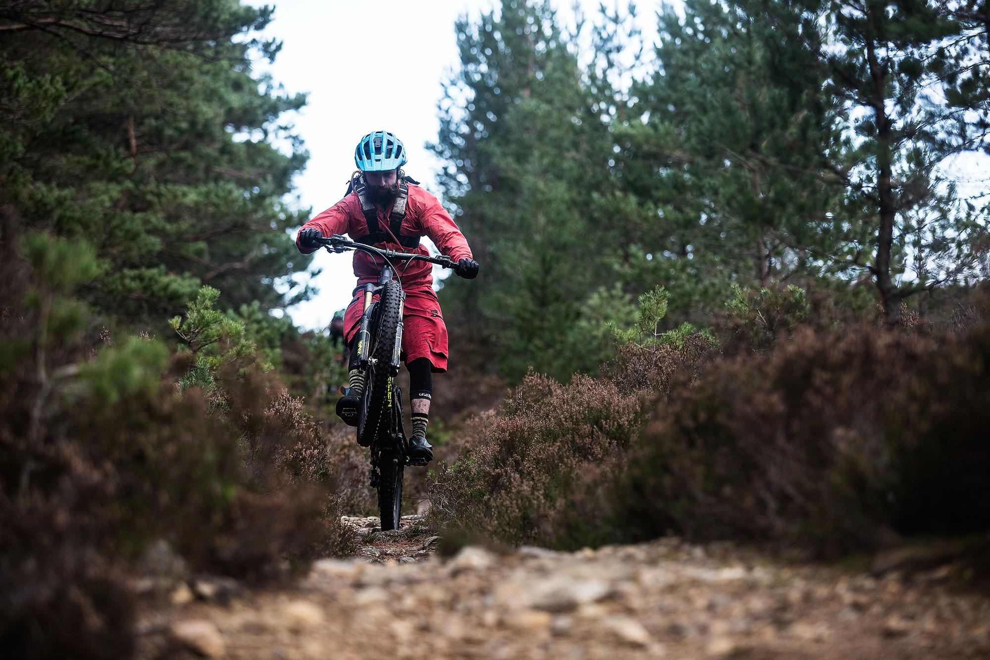 A mountain biker wearing a blue helmet and a red outfit rides a bike along a rocky trail surrounded by green trees and shrubs. The biker is performing a wheelie, lifting the front wheel off the ground as they navigate the path. Rocky Road mountain bike trail.