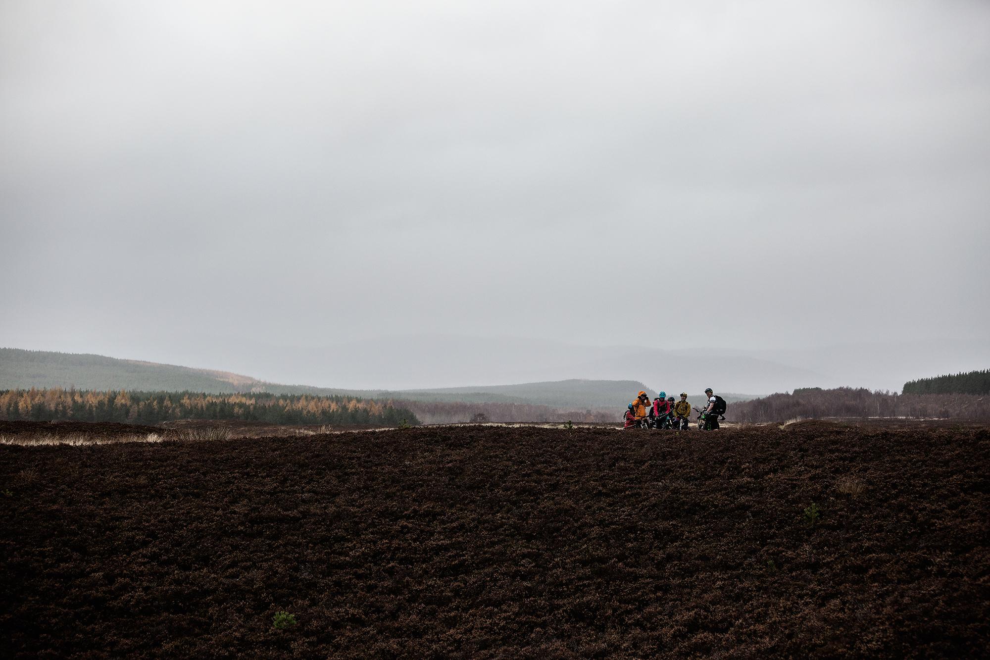 A group of people sitting on a grassy area with a view of rolling hills and overcast skies in the background. The landscape features a mix of sparse vegetation and forested areas, set against a muted, cloudy sky. River Feshie Trail mountain bike trail.