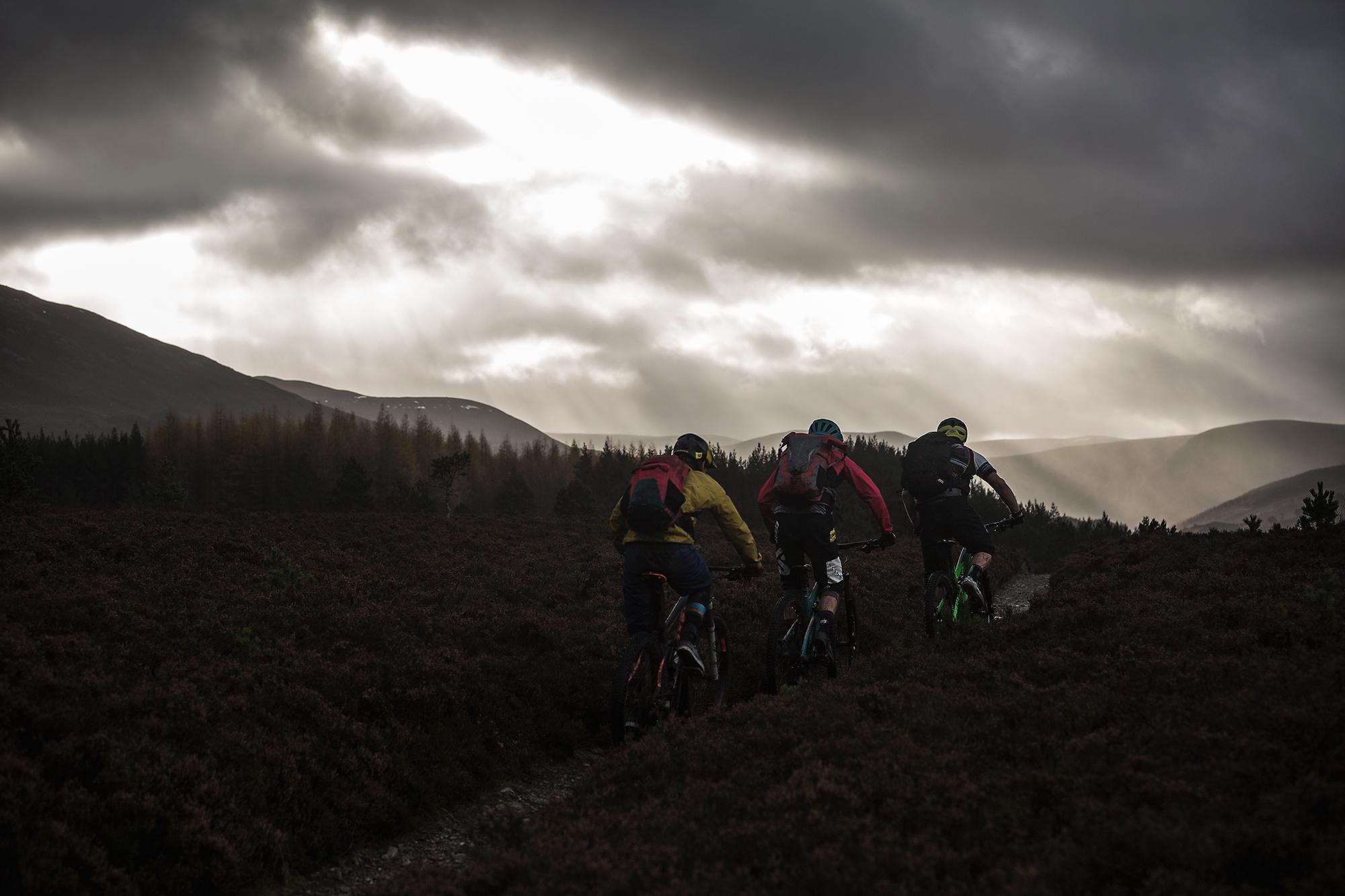 Four mountain bikers riding along a trail through a moorland landscape under a dramatic, cloudy sky. The setting features rolling hills in the background, with sunlight breaking through the clouds, casting rays onto the terrain. The cyclists are dressed in colorful outdoor gear and are seen from behind as they navigate the path. River Feshie Trail mountain bike trail.