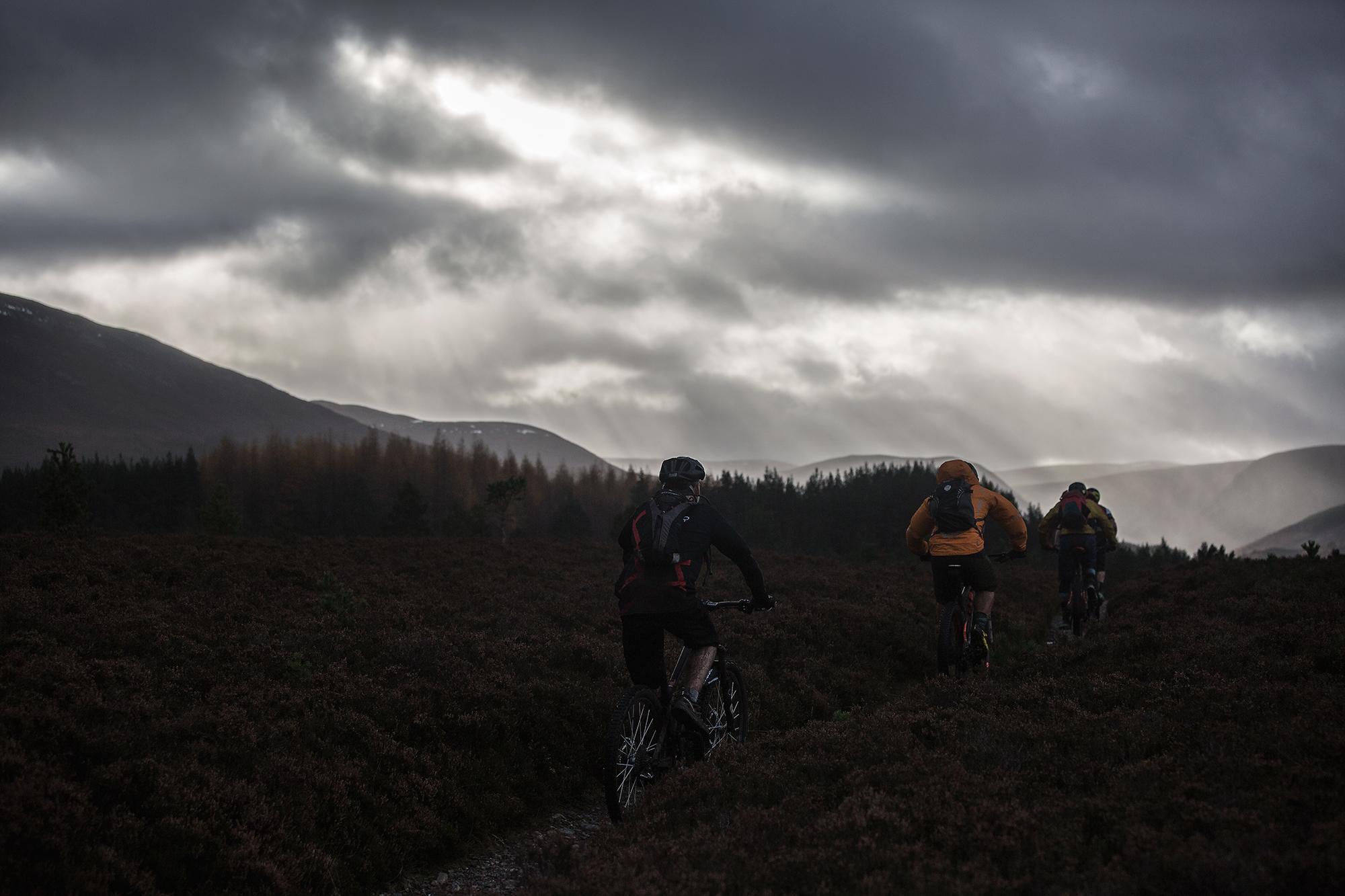 A group of mountain bikers riding along a trail in a grassy landscape under a dramatic, cloudy sky, with mountains in the background. River Feshie Trail mountain bike trail.