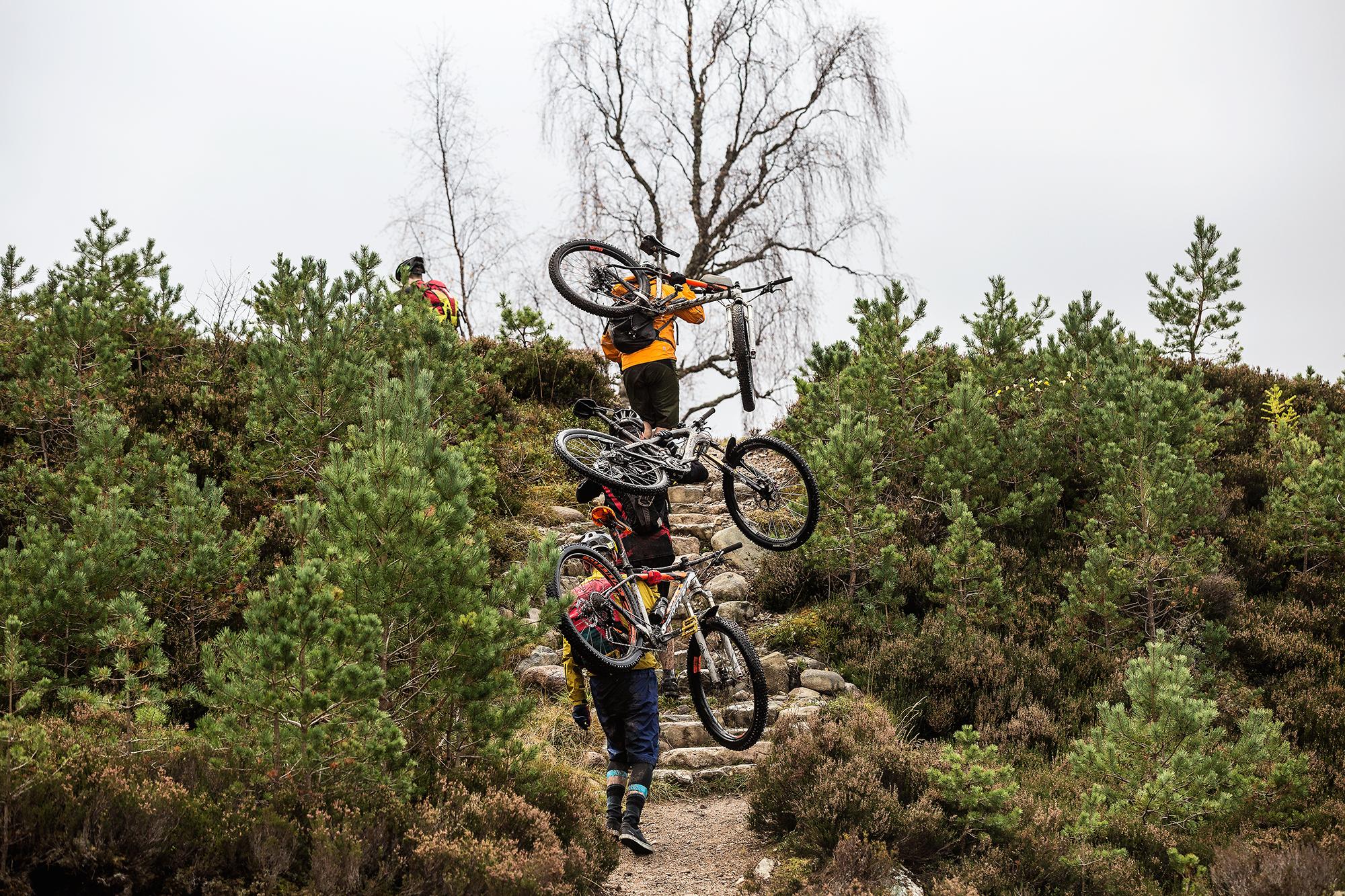A group of mountain bikers navigating a rocky path through a wooded area, carrying their bikes as they ascend a steep incline. The scene captures the rugged terrain and greenery surrounding them, with a cloudy sky overhead. River Feshie Trail mountain bike trail.