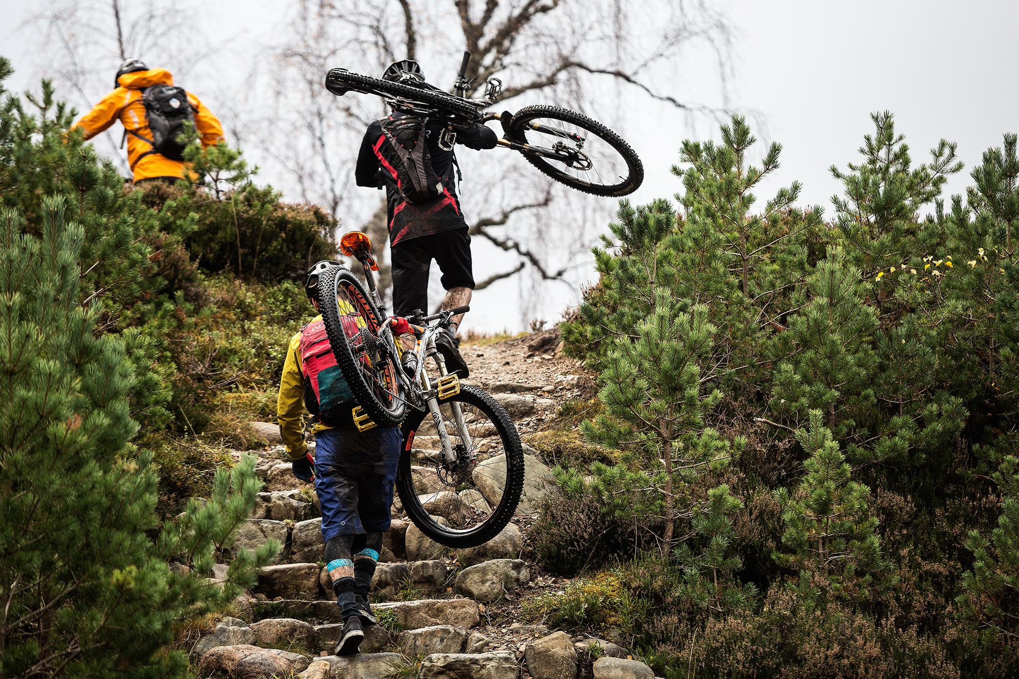 Two mountain bikers are climbing a rocky path surrounded by greenery, with one biker carrying a bike over their shoulder. The scene features a rugged outdoor environment, and both riders are dressed in colorful, athletic gear suited for biking. The sky appears cloudy, suggesting a cool atmosphere. River Feshie Trail mountain bike trail.