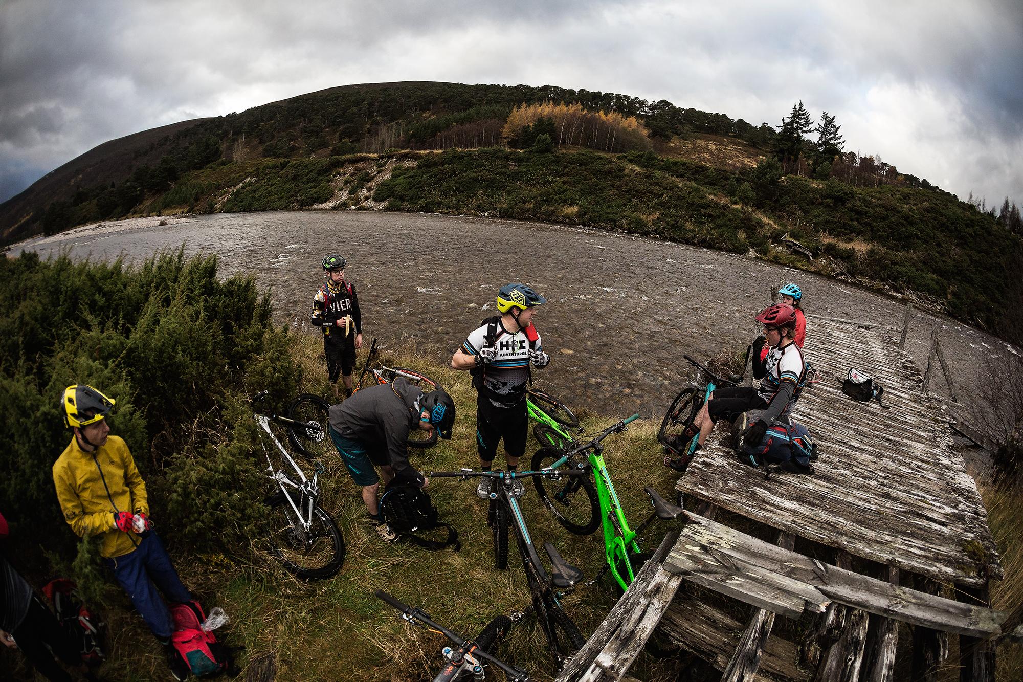 A group of mountain bikers gathered near a riverbank, with several bicycles positioned around them. Some cyclists are inspecting their gear while others are taking a break by a wooden platform. The backdrop features green hills under a cloudy sky, indicating an adventurous outdoor setting. River Feshie Trail mountain bike trail.