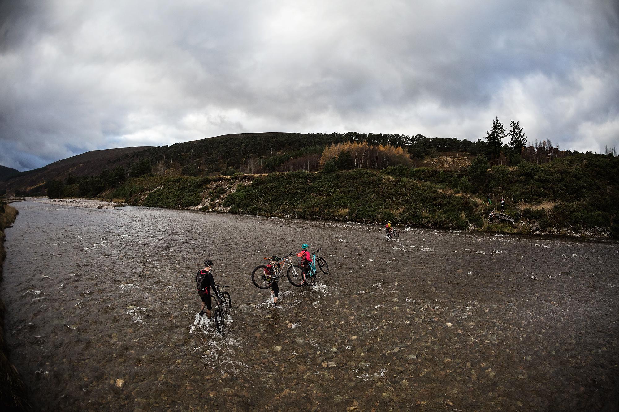 Four cyclists are crossing a shallow river, carrying their bikes above the water. The landscape features rolling hills and a mix of greenery, with a cloudy sky overhead. The scene captures a sense of adventure and outdoor activity in a natural setting. River Feshie Trail mountain bike trail.
