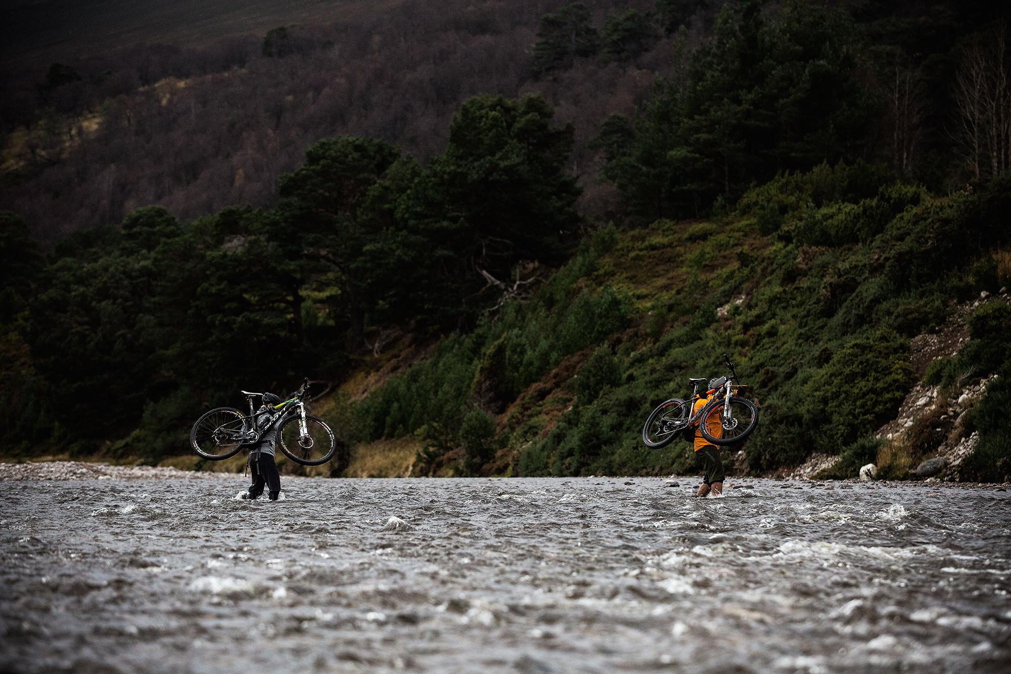 Two individuals wade through a river while carrying mountain bikes on their shoulders. The surrounding landscape features lush greenery and steep, wooded hills in the background. The scene captures the adventurous spirit of outdoor cycling in a natural setting. River Feshie Trail mountain bike trail.