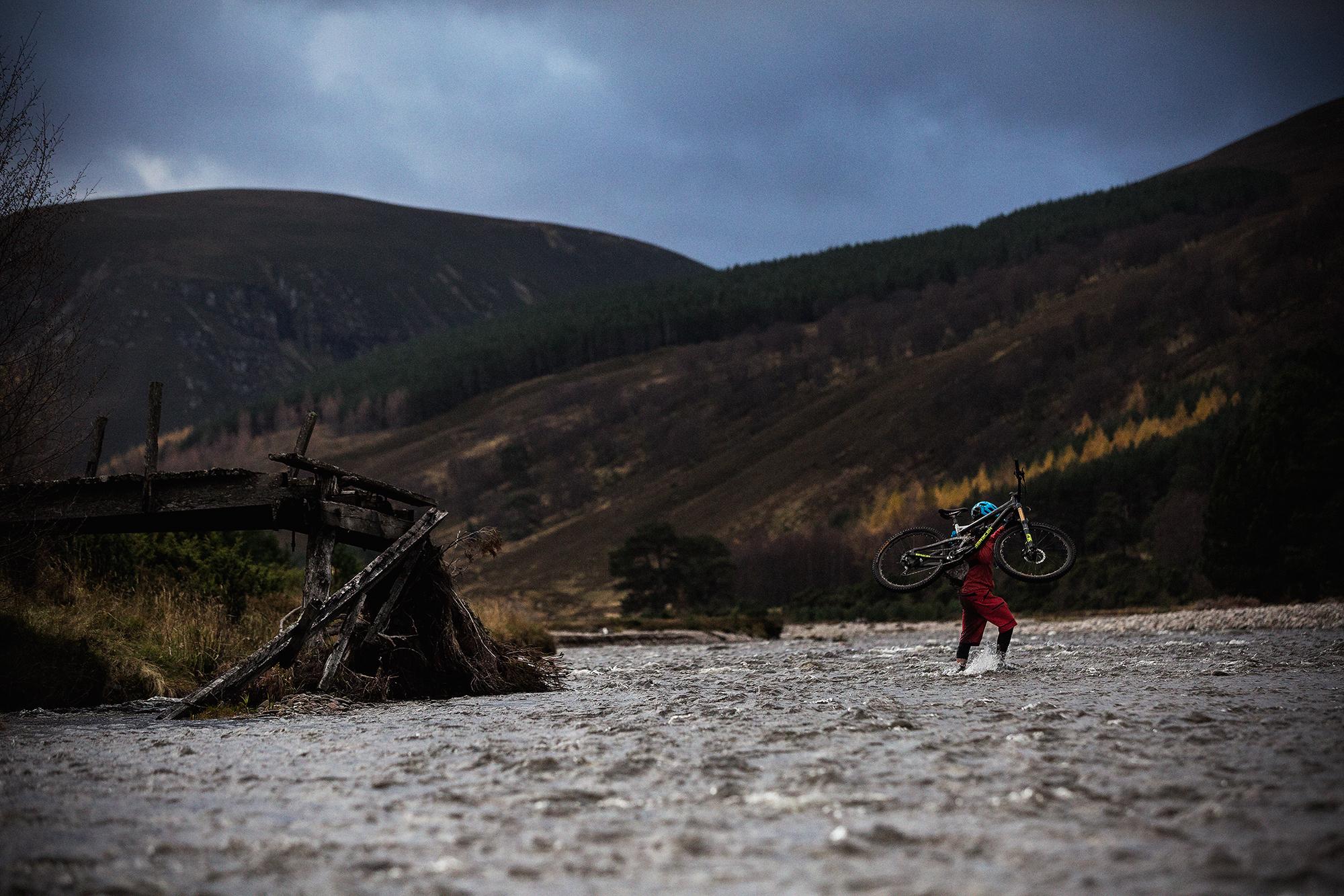 A person wades through a river while carrying a mountain bike over their shoulder. The scene is set against a backdrop of rolling hills and a partially cloudy sky, with a dilapidated wooden structure visible in the foreground. The landscape features trees and grassy areas, suggesting a remote and rugged environment. River Feshie Trail mountain bike trail.