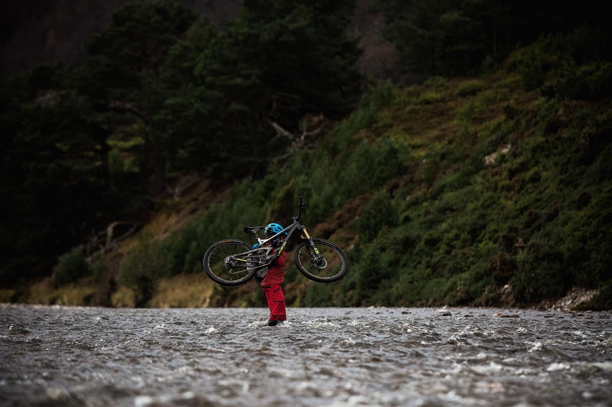 A person dressed in red is standing in a shallow, flowing river while holding a mountain bike above their head. Lush green hills and a line of trees are visible in the background, suggesting an outdoor adventure setting. The scene captures a moment of triumph or challenge in a rugged natural environment. River Feshie Trail mountain bike trail.