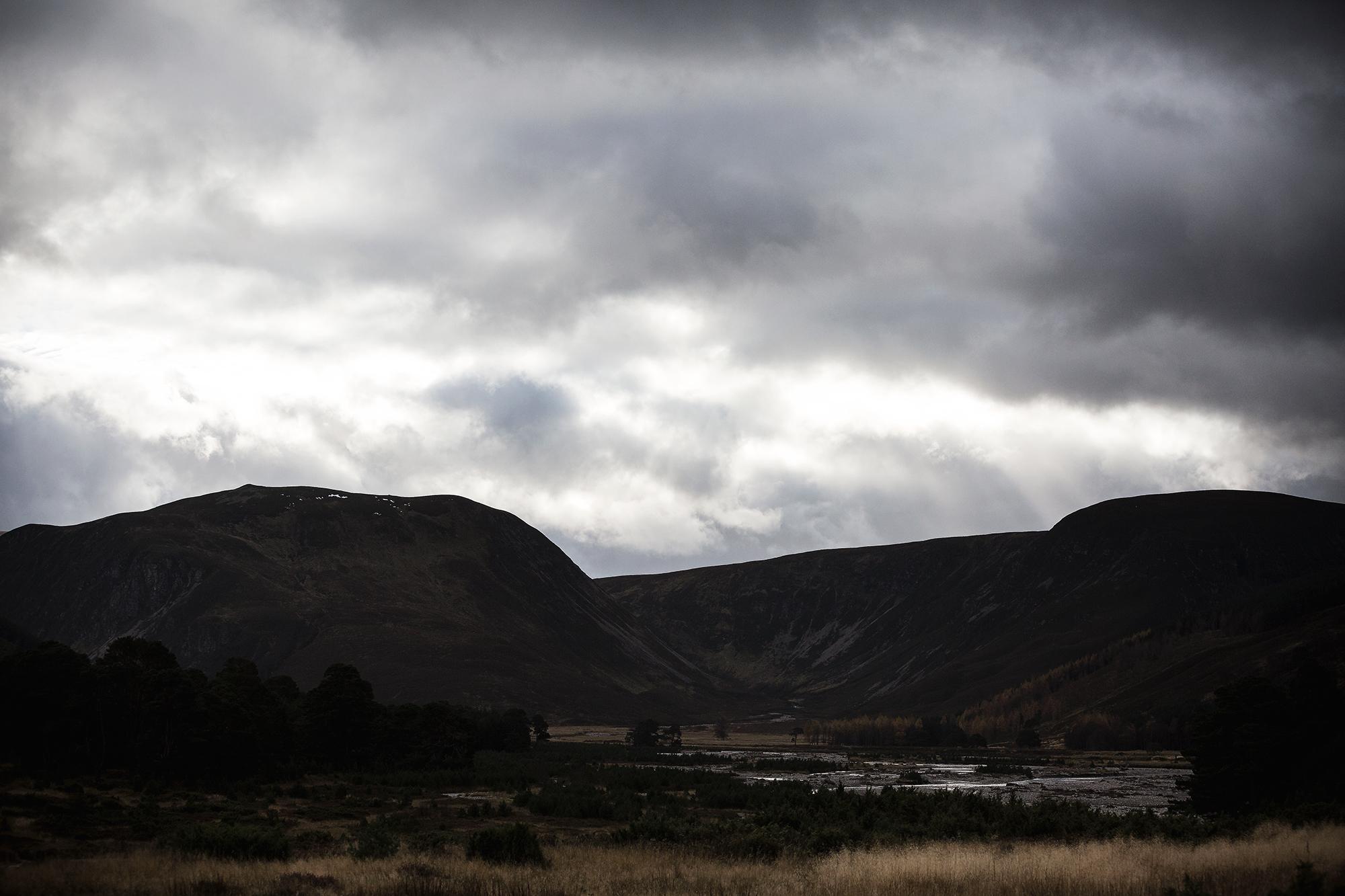 A dramatic landscape featuring rolling hills and mountains under a cloudy sky. The foreground includes patches of grass and sparse trees, while a river can be seen meandering through the valley. The atmosphere is moody and atmospheric, with hints of light breaking through the clouds. River Feshie Trail mountain bike trail.