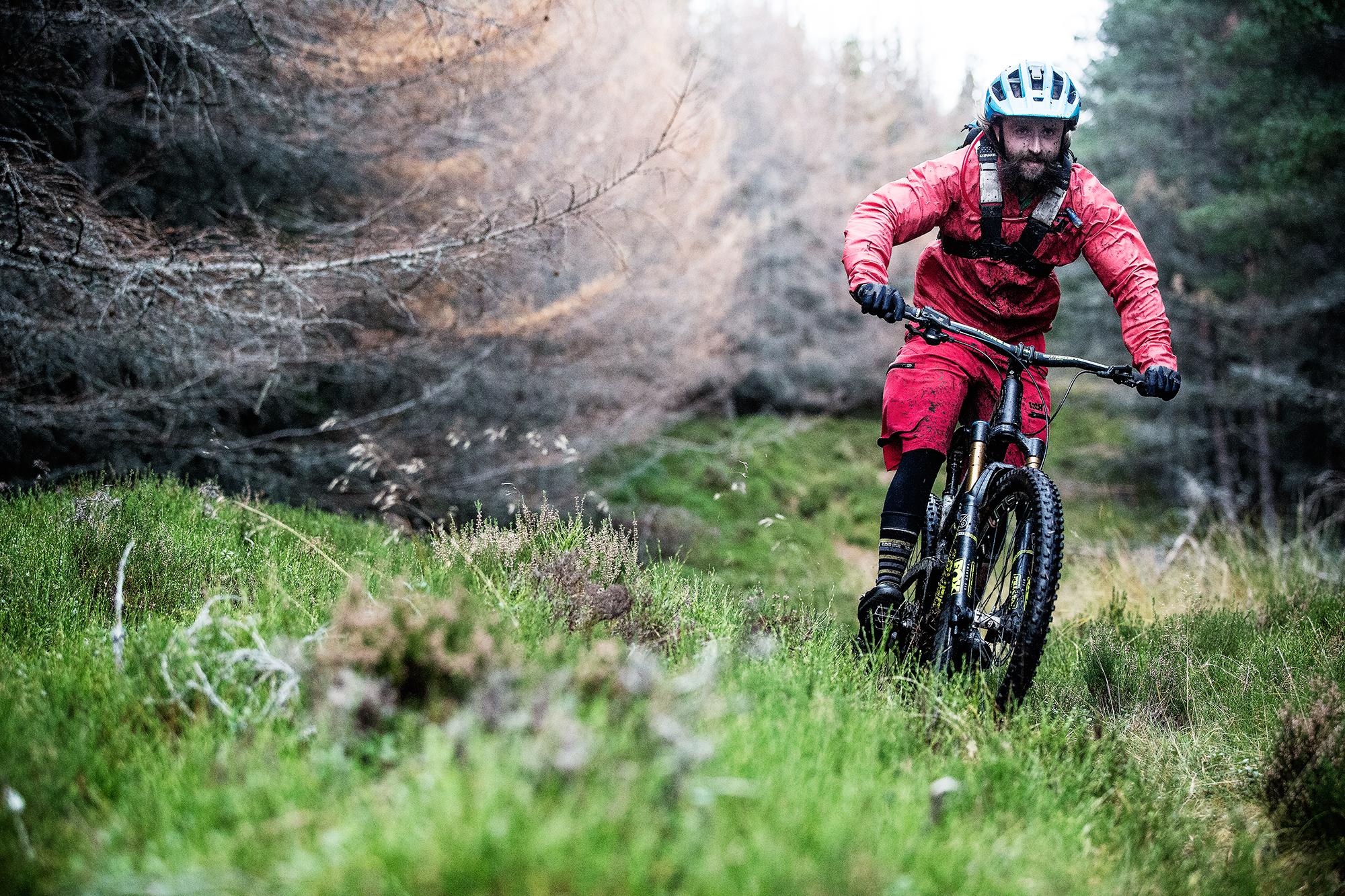 A mountain biker in a red outfit and helmet rides aggressively through a lush, green forest trail, surrounded by trees and underbrush. The scene captures the excitement of outdoor biking in a natural setting. The Lodge Descent mountain bike trail.