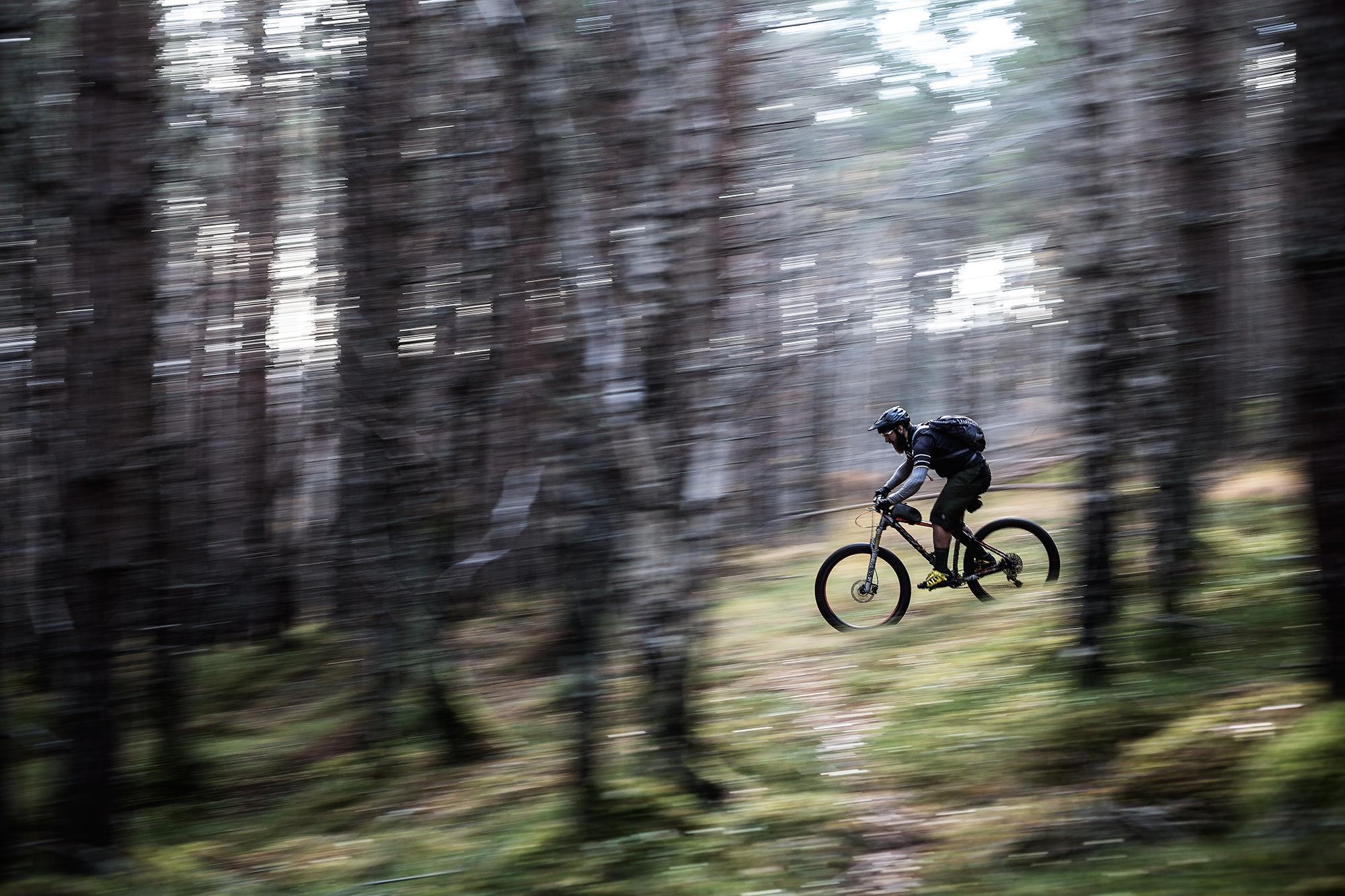 A mountain biker riding quickly along a narrow trail through a dense forest, with trees blurred in the background to convey motion. The biker is wearing a helmet and backpack, navigating the winding path in a natural setting. Green Dream mountain bike trail.