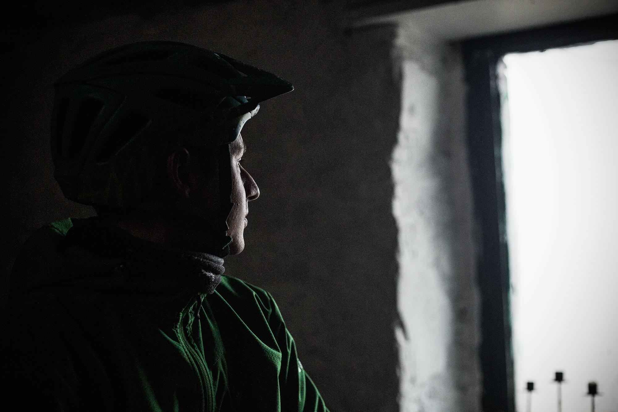 A person wearing a green cycling helmet and jacket is seen in profile, gazing thoughtfully out of a window. The dimly lit interior contrasts with the bright light coming from the window, creating a dramatic atmosphere. Rhyvoan Pass mountain bike trail.