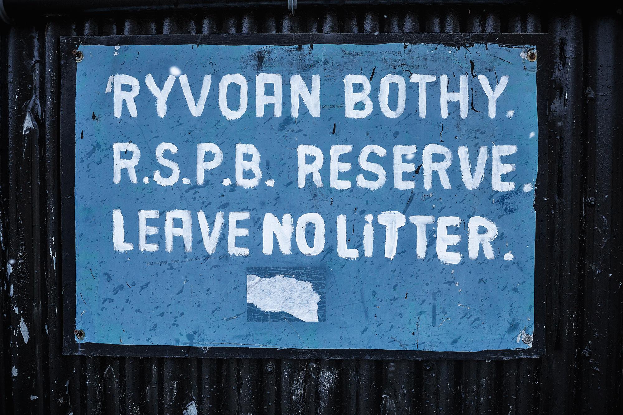 Signboard at Ryvoan Bothy, R.S.P.B. Reserve, with the message "Leave No Litter" painted in white on a blue background. The sign is weathered, with some peeling paint, and is mounted on a black, textured surface. Rhyvoan Pass mountain bike trail.