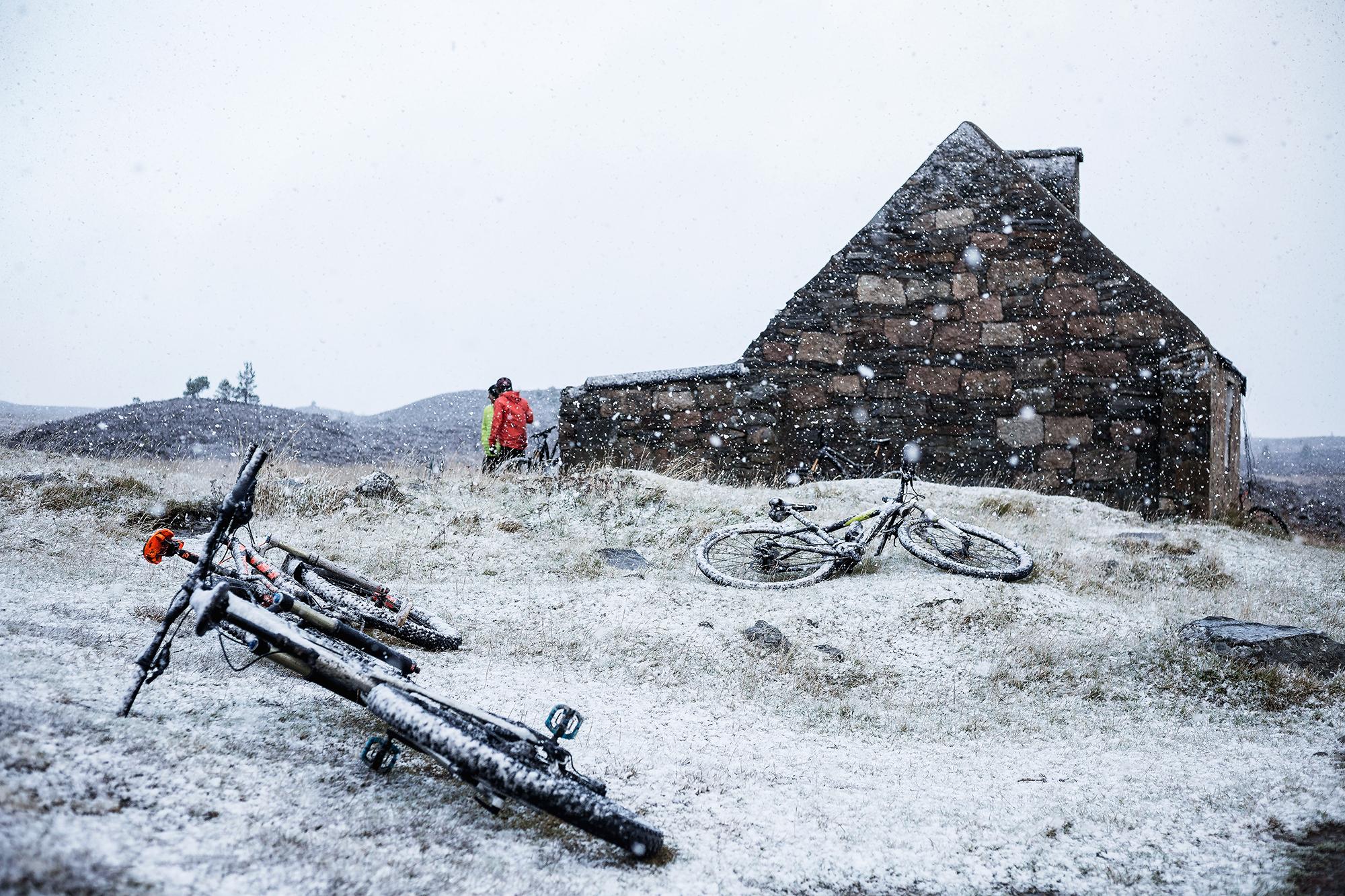 A snowy landscape featuring two bicycles lying on the ground near an old stone building. In the background, a person in a bright red jacket is seen approaching the building, with snow falling around them. The terrain is grassy with patches of snow, and rolling hills can be seen in the distance under a gray sky. Rhyvoan Pass mountain bike trail.